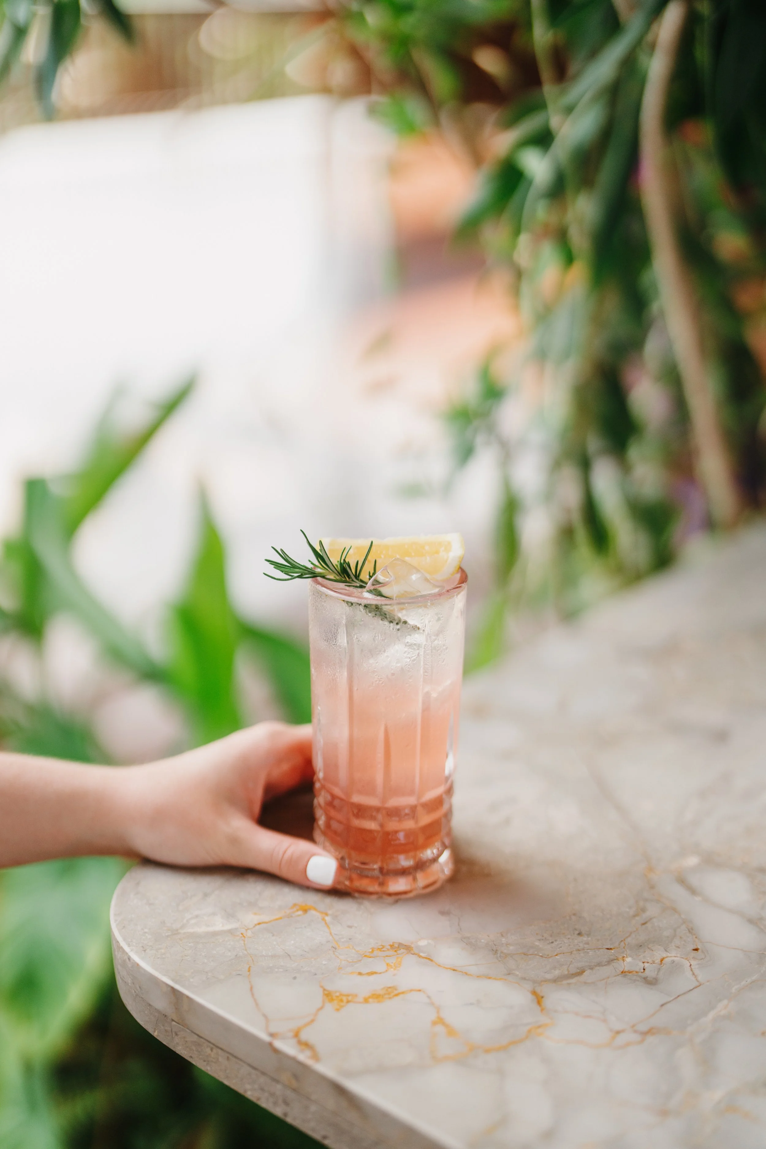 A hand holding a tall glass of pink cocktail garnished with lemon wedge and rosemary sprig on a marble table.
