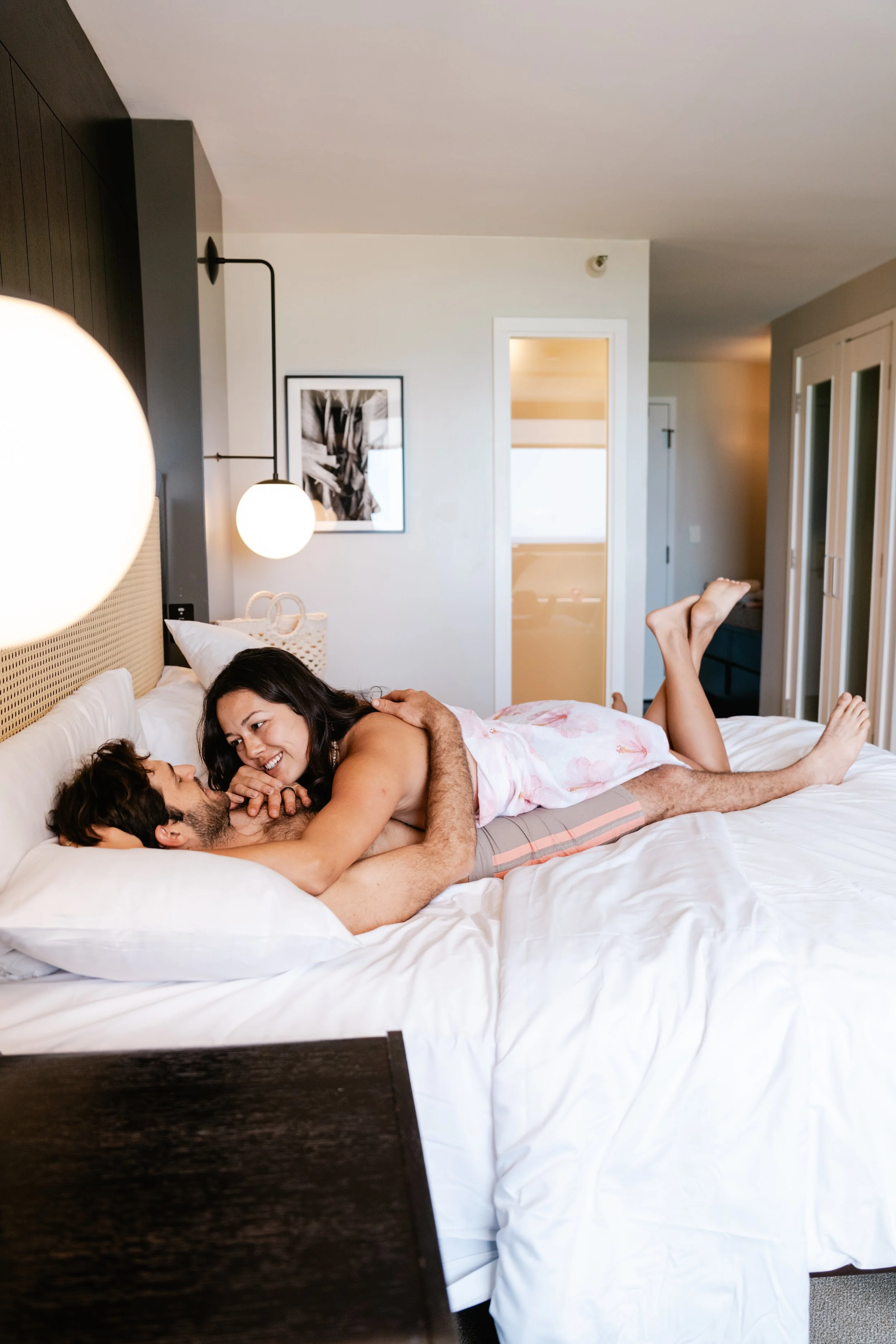 A woman and a man playfully cuddling on a bed in a modern bedroom, smiling and looking at each other.