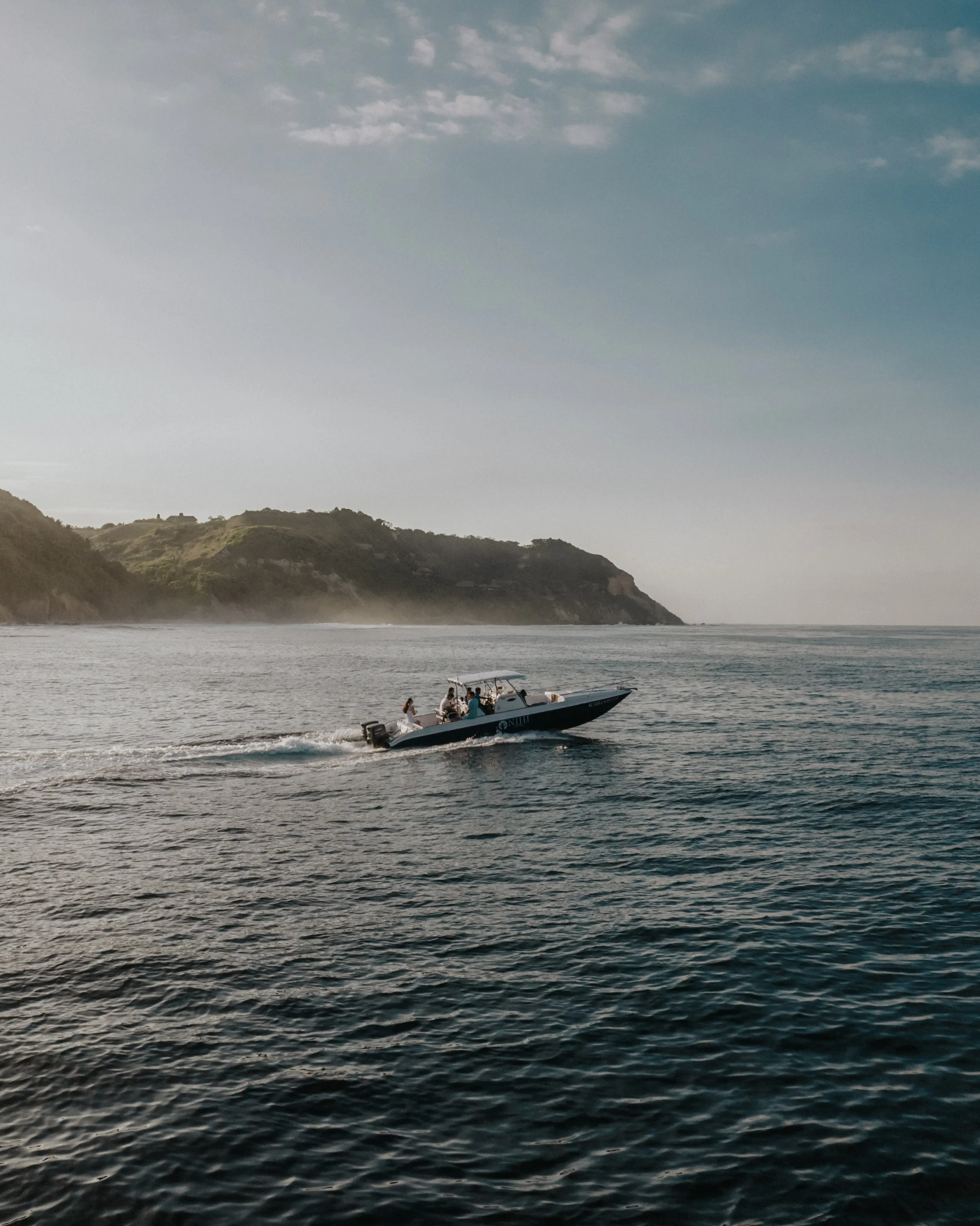 A boat moving through the water near a shoreline with green hills and a slightly cloudy sky.