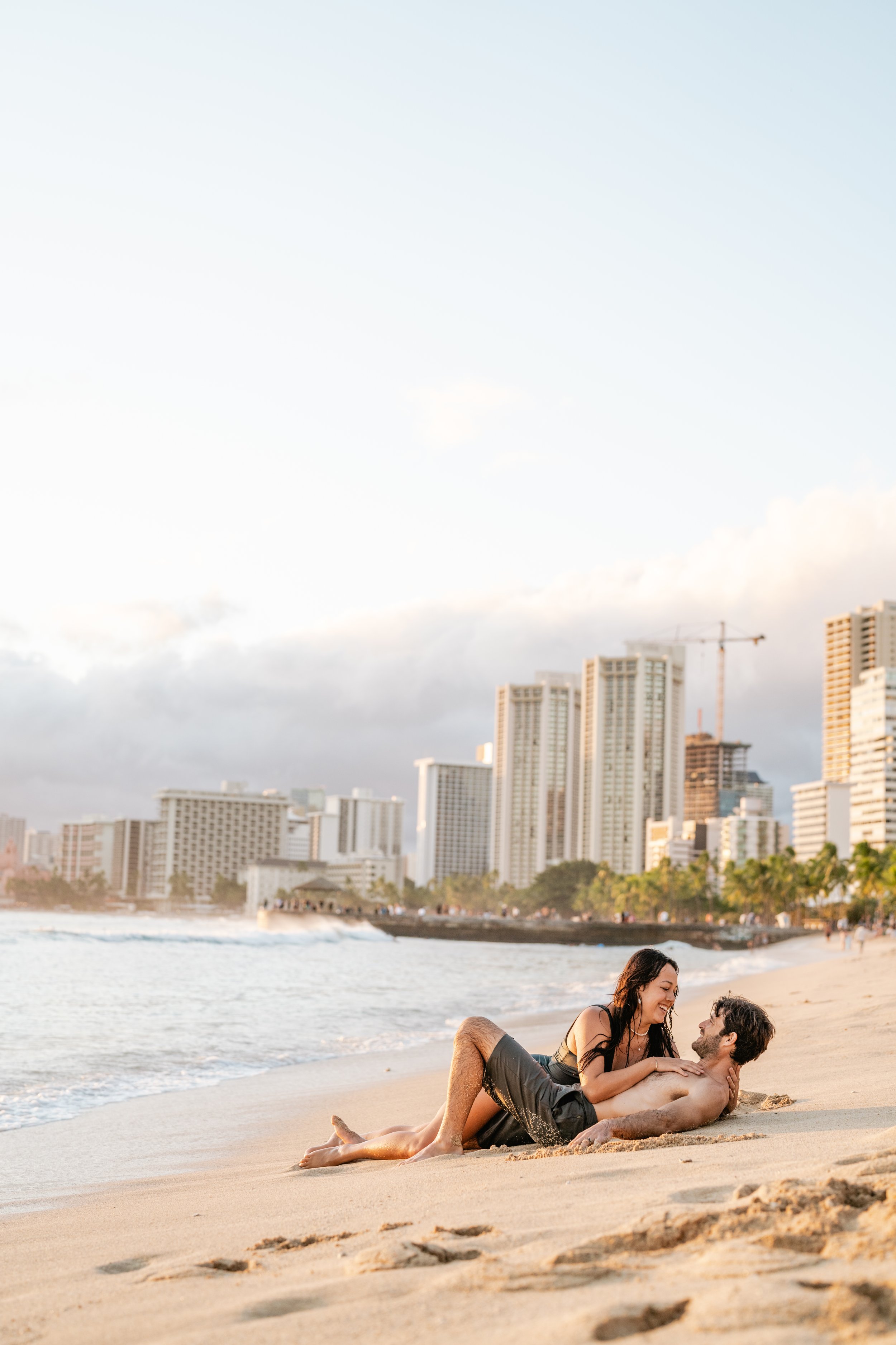 A couple is relaxing on the sandy beach with a city skyline in the background, possibly in Hawaii or a tropical location.