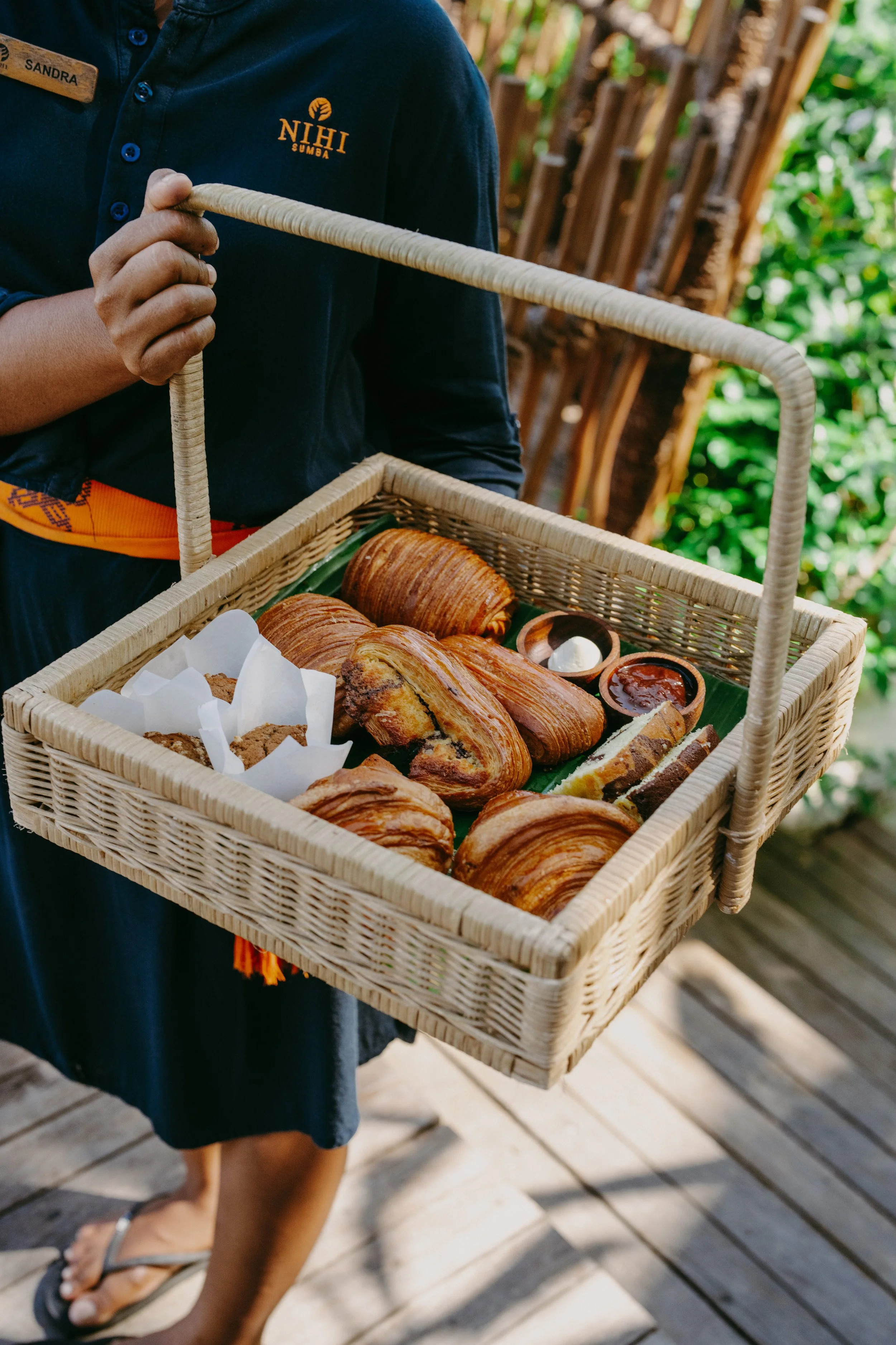 Person holding a basket of assorted pastries and condiments, wearing a navy shirt with a name tag and a logo, standing on wooden decking.