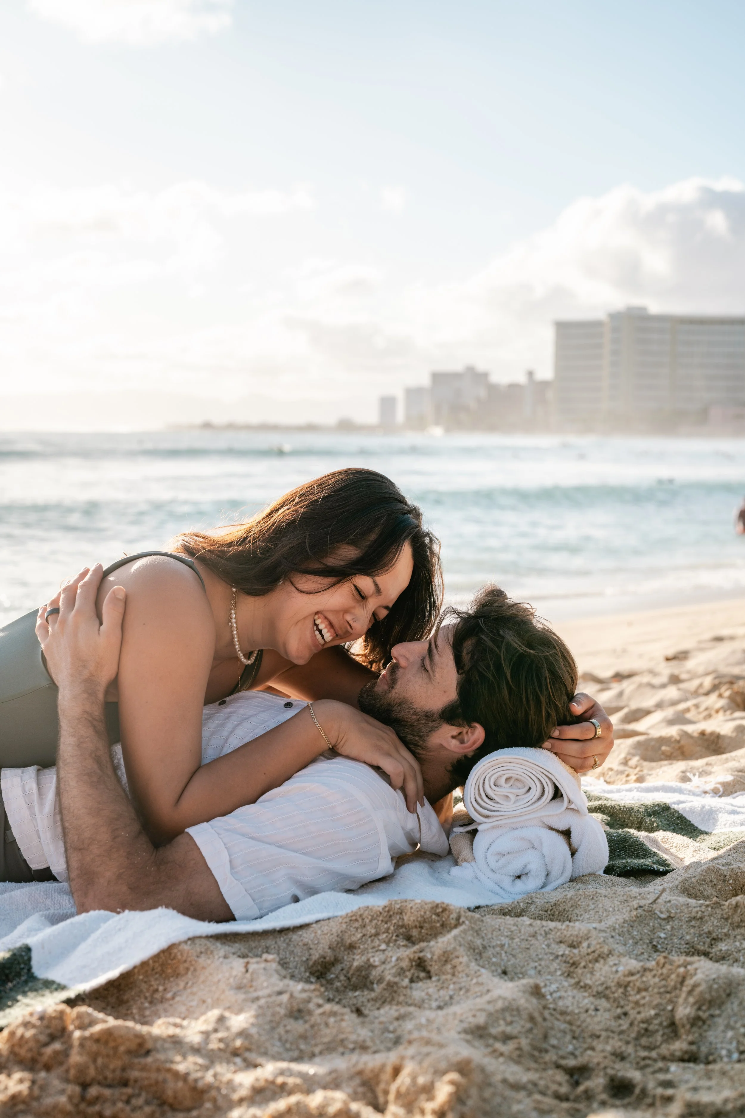 A happy couple lying on the beach, smiling and cuddling, with the ocean and cityscape in the background.