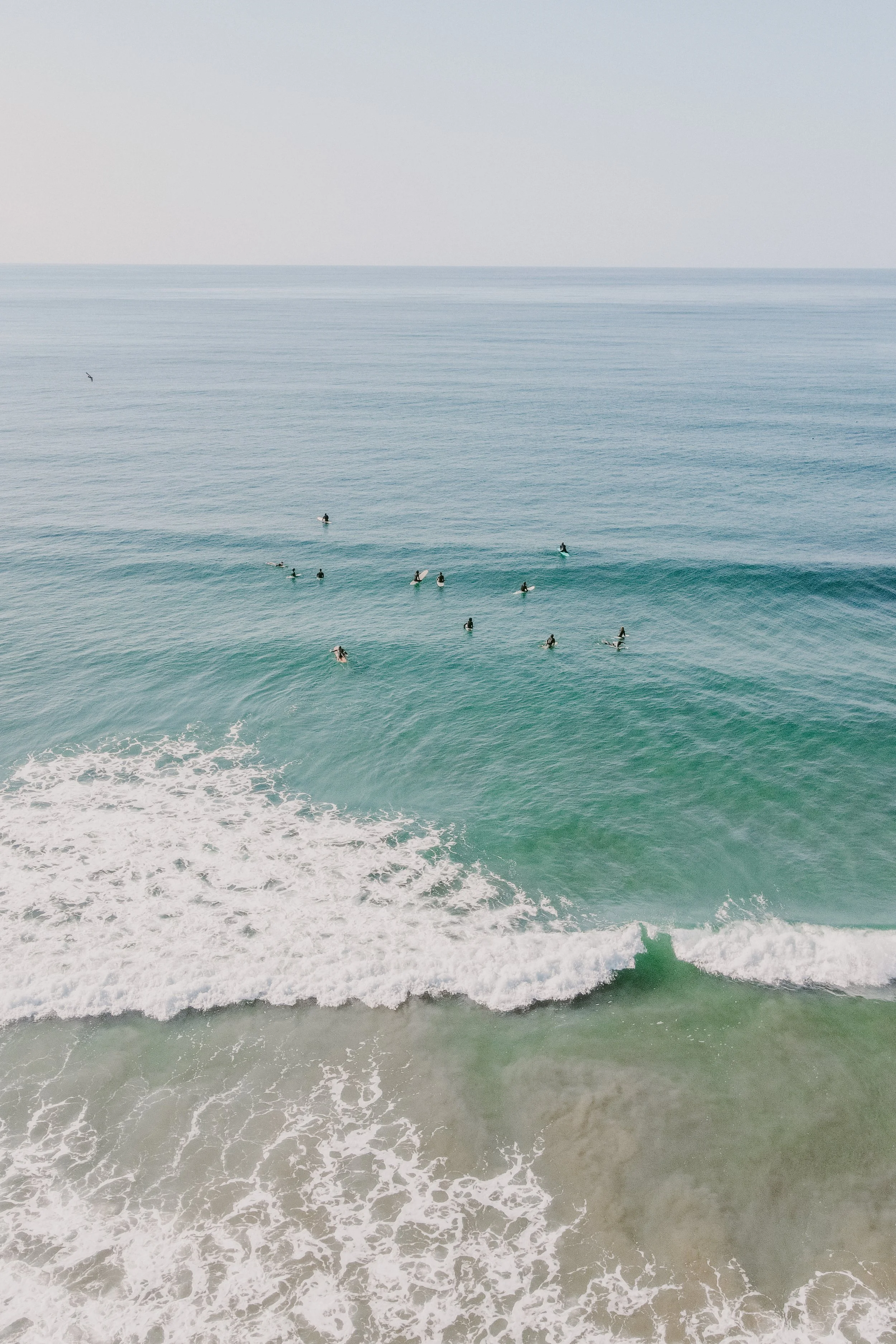 Aerial view of the ocean with surfers waiting on their surfboards near the shoreline.