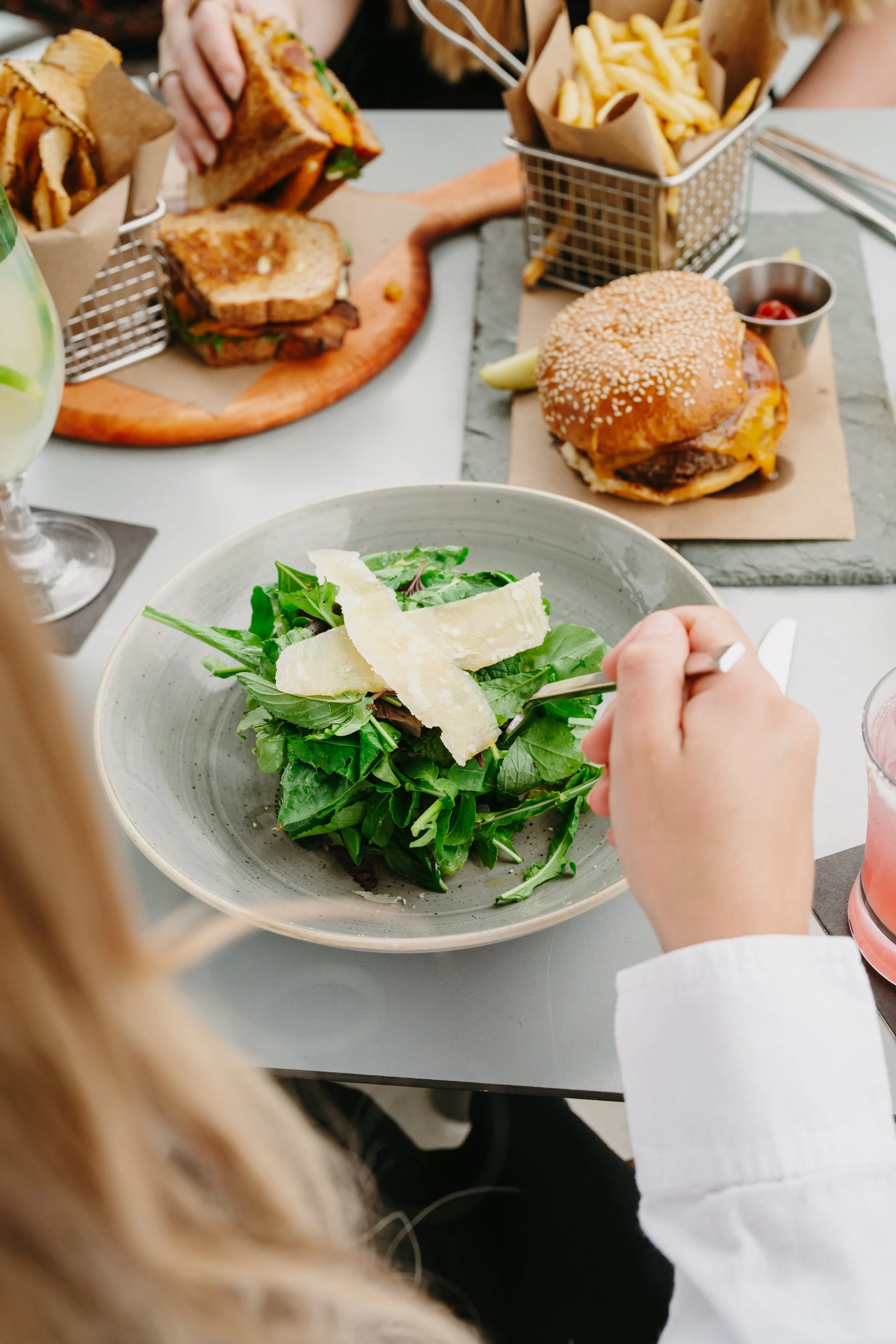A person dining at a table with a green salad topped with cheese in a gray bowl, a cheeseburger with a sesame seed bun, lettuce, tomato, and a side of french fries, along with a sandwich with bacon, all arranged on the table.