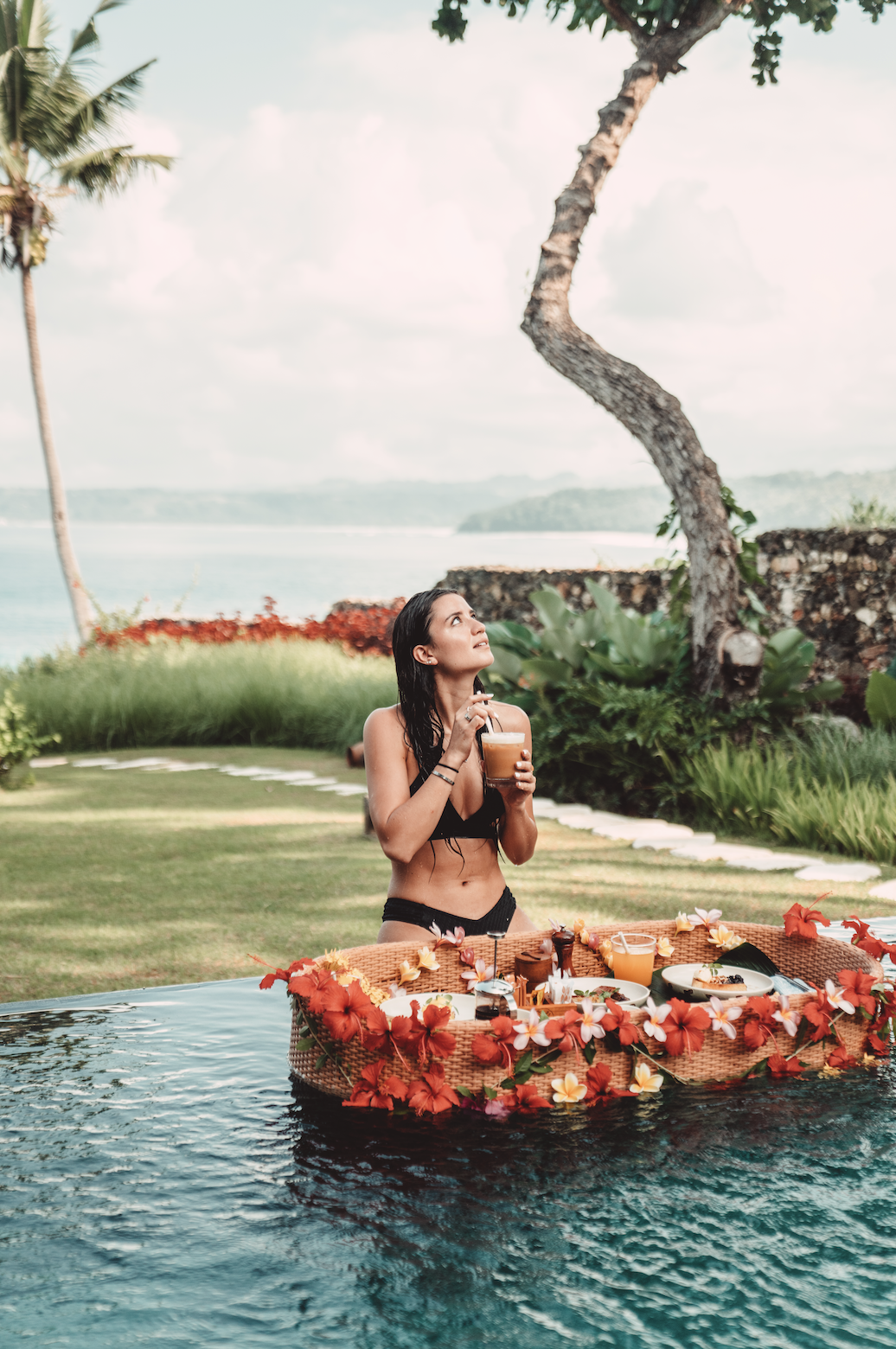 A woman in a black bikini standing in a swimming pool, holding a drink, with a floating tray of breakfast decorated with flowers, in a tropical outdoor setting near water and palm trees.