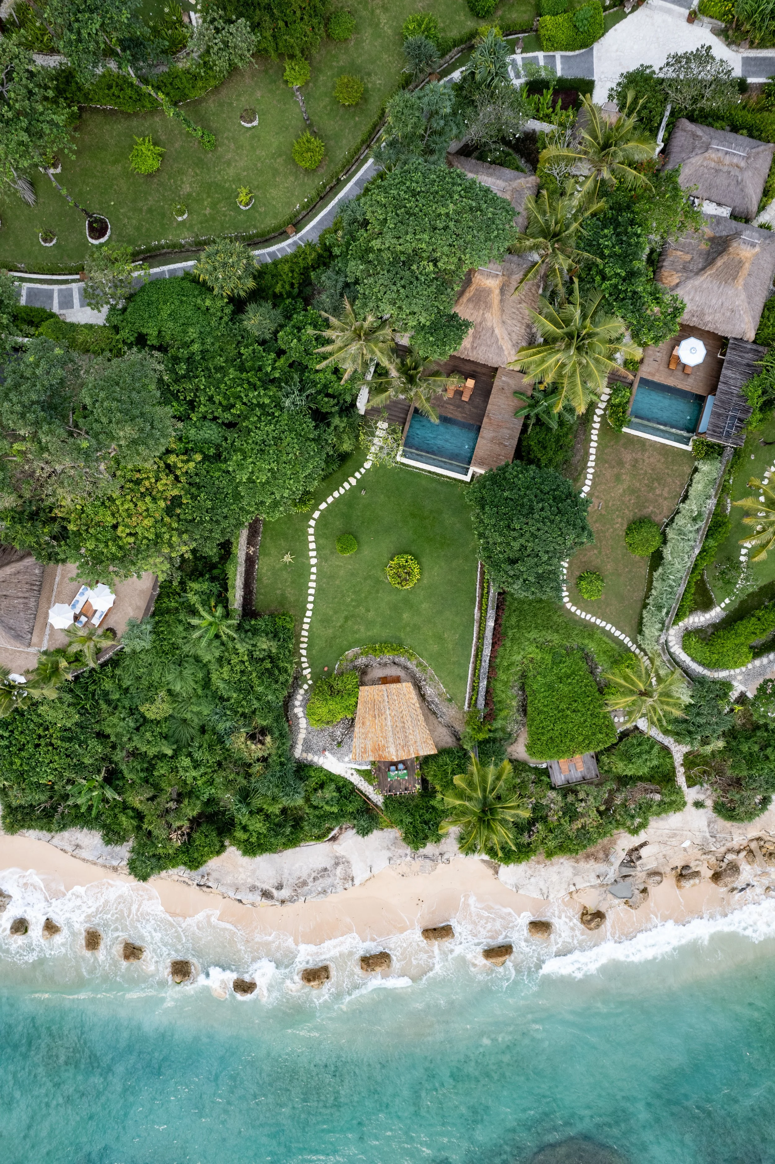 Aerial view of a tropical resort with green lawns, trees, thatched roof bungalows, swimming pools, walkways, and a beach with waves and rocks.