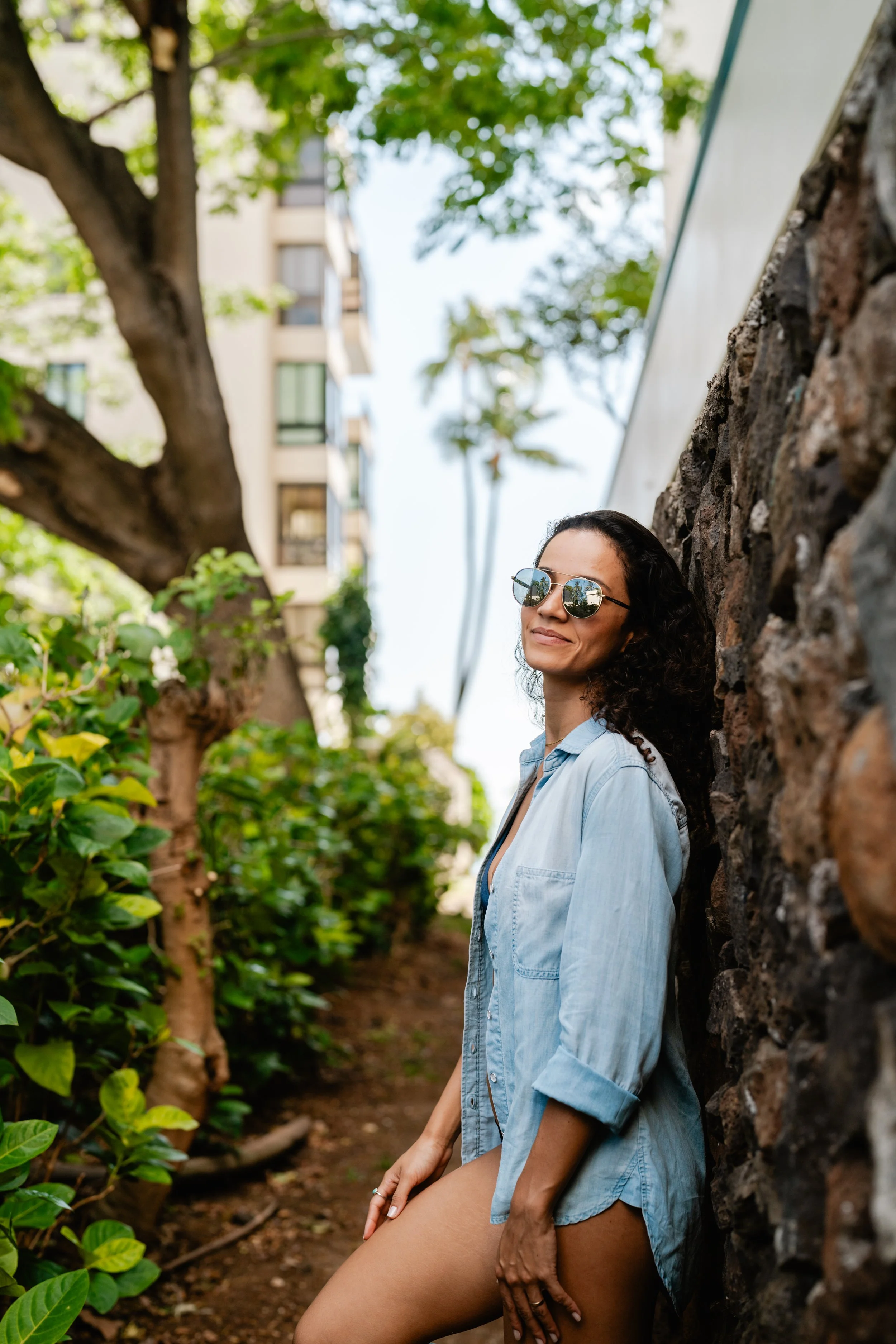 A woman in sunglasses and a denim shirt leaning against a stone wall outdoors surrounded by green plants and trees, with buildings visible in the background.