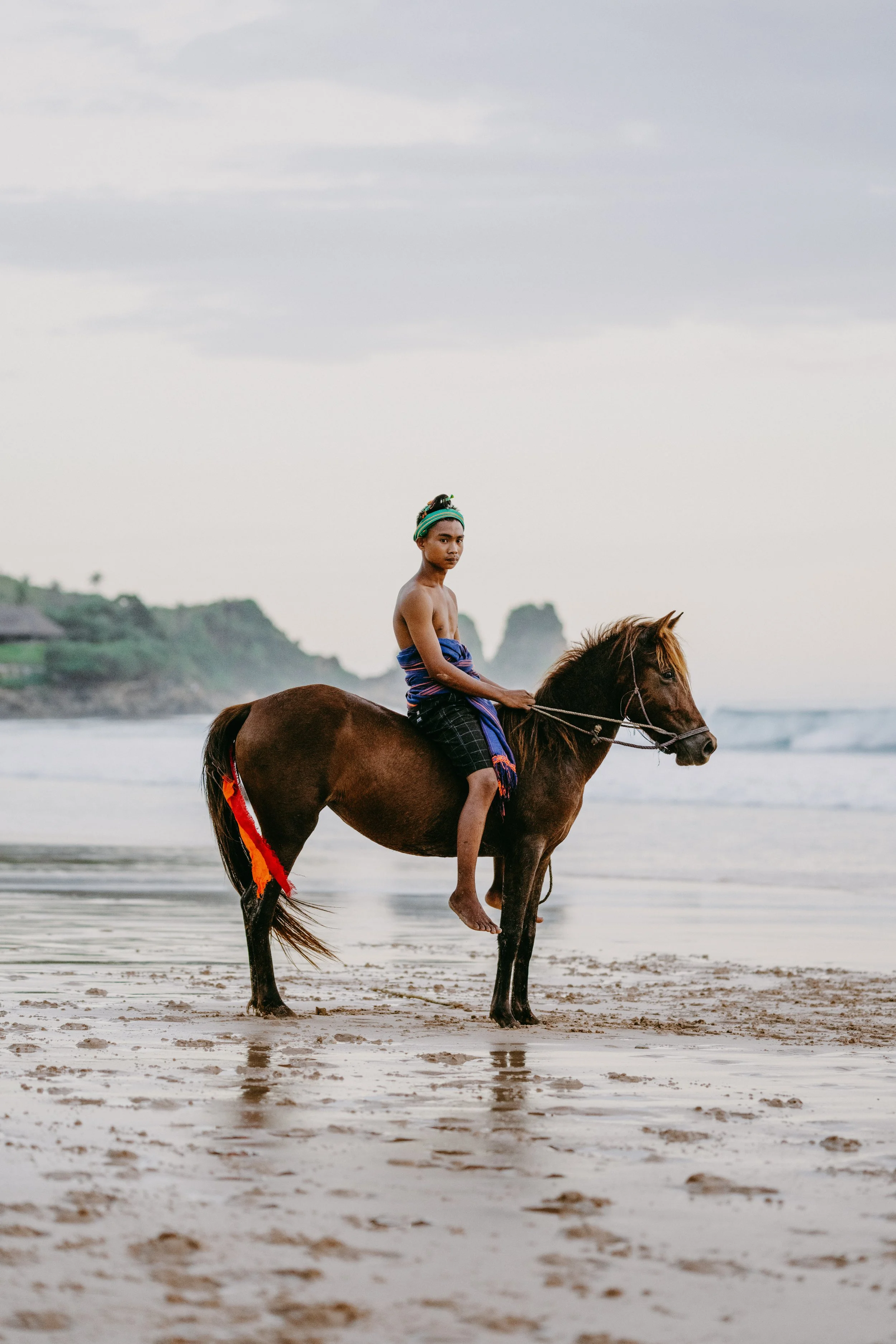 A young man riding a horse on the beach with cliffs and ocean in the background.