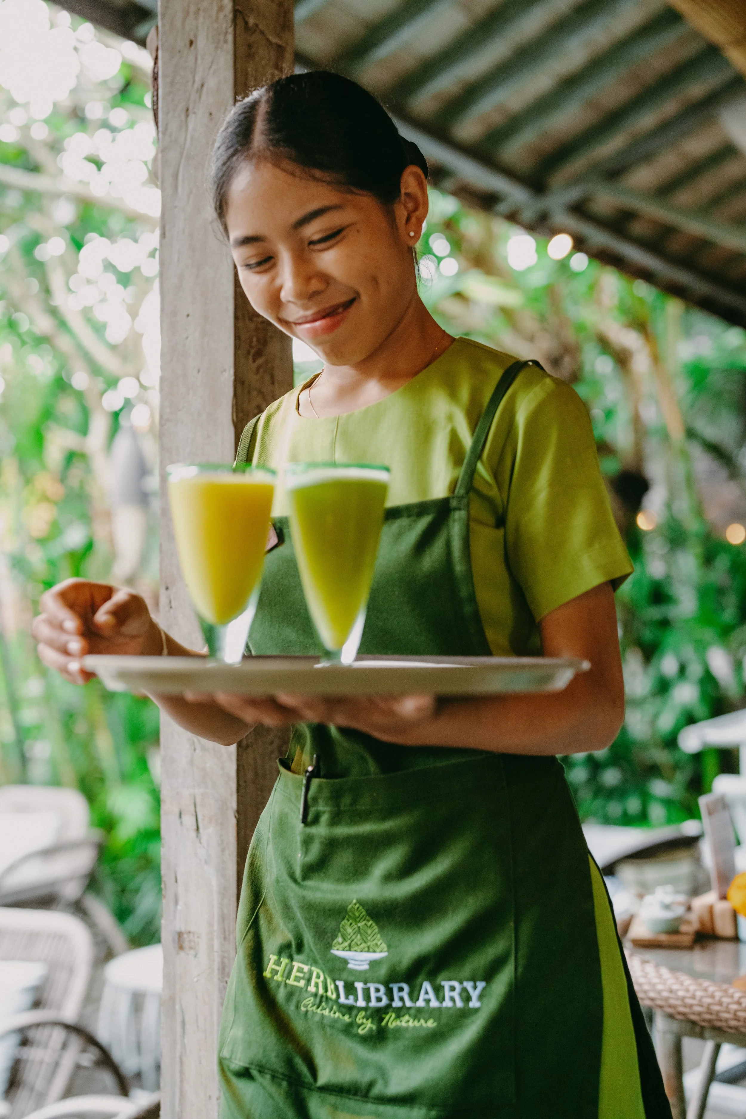 A woman in a green apron holding a tray with two glasses of green juice, smiling in an outdoor setting with trees and a thatched roof in the background.
