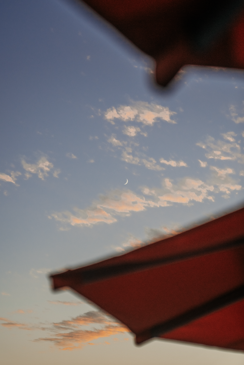 Sky with pink clouds, crescent moon, viewed through a window or opening with red structure at the top and bottom.