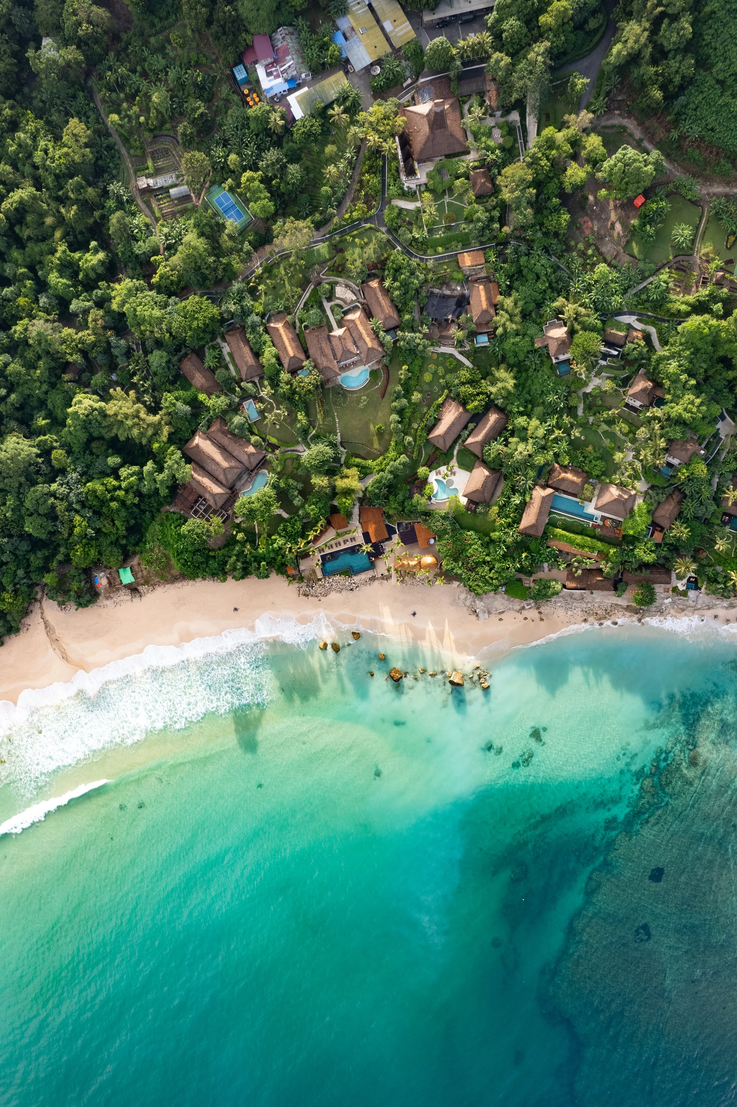 Aerial view of a beachside resort with multiple villas, swimming pools, lush green trees, and a sandy beach with turquoise waters.