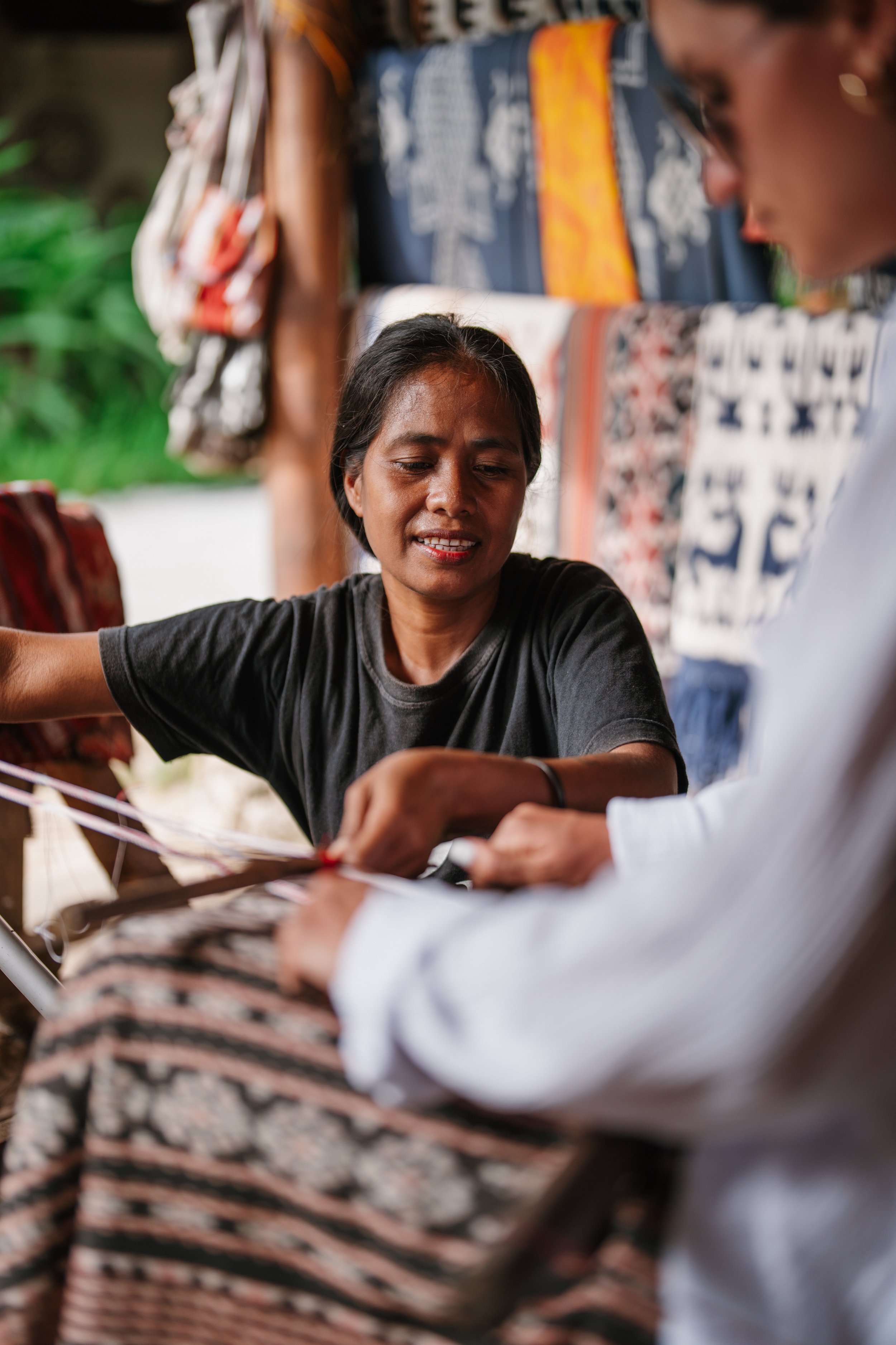 Two women are engaged in a craft or textile activity, with woven fabrics hanging behind them, in an outdoor setting with greenery in the background.