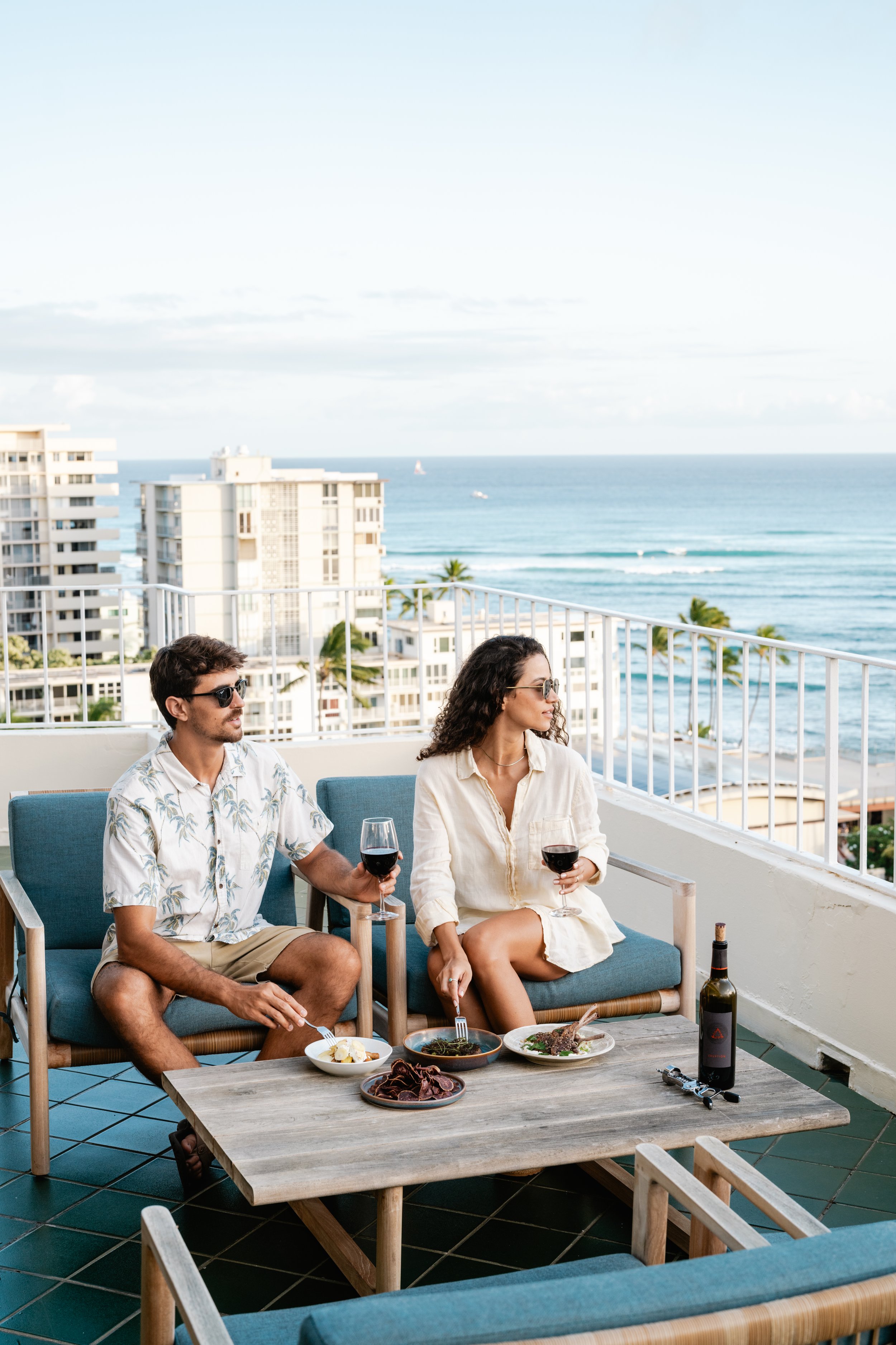 A man and woman enjoying a meal and wine on a balcony overlooking the ocean and city buildings.