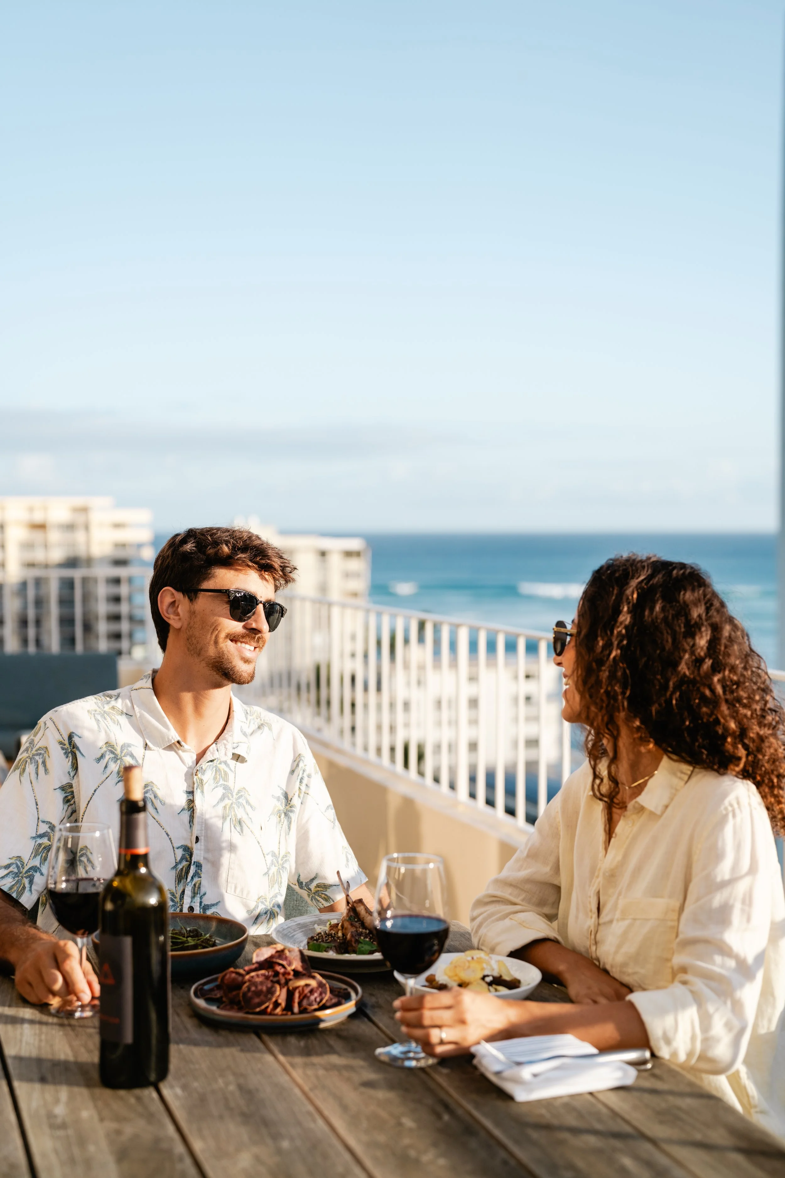 A man and woman enjoying a meal and conversation on a balcony with ocean view, with wine and food on the table.