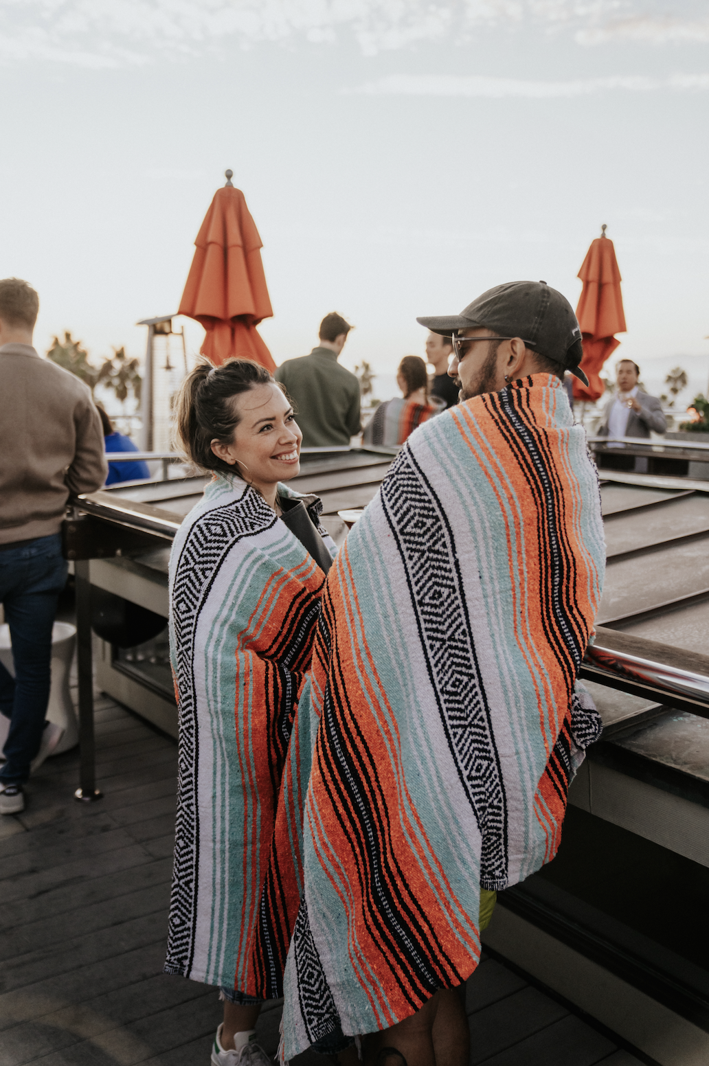 Two people wrapped in colorful striped blankets talking on a rooftop bar with orange umbrellas and other patrons in the background.