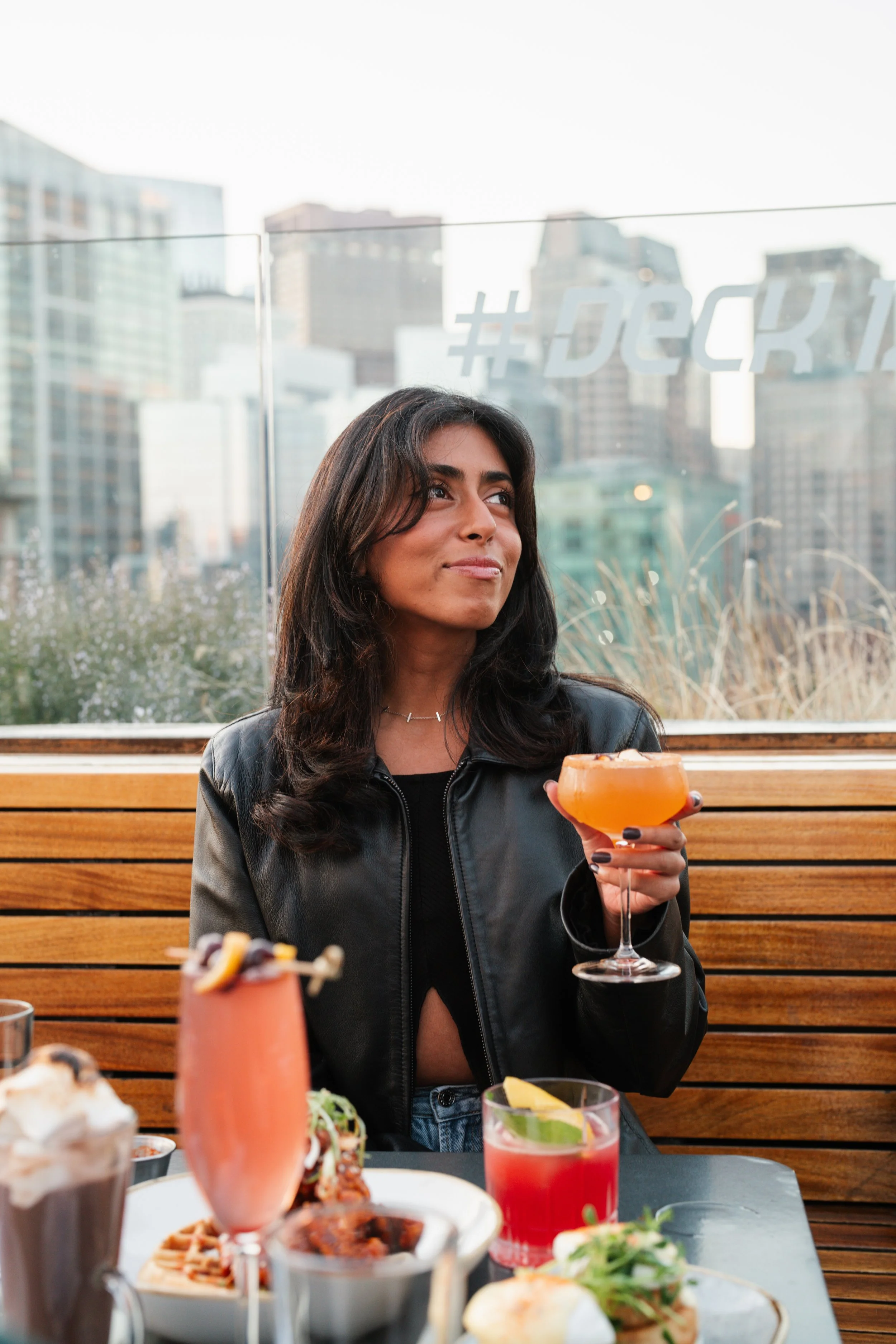 A woman with dark hair sitting at a rooftop restaurant holding a pink cocktail, surrounded by other colorful drinks and food, against a cityscape background.
