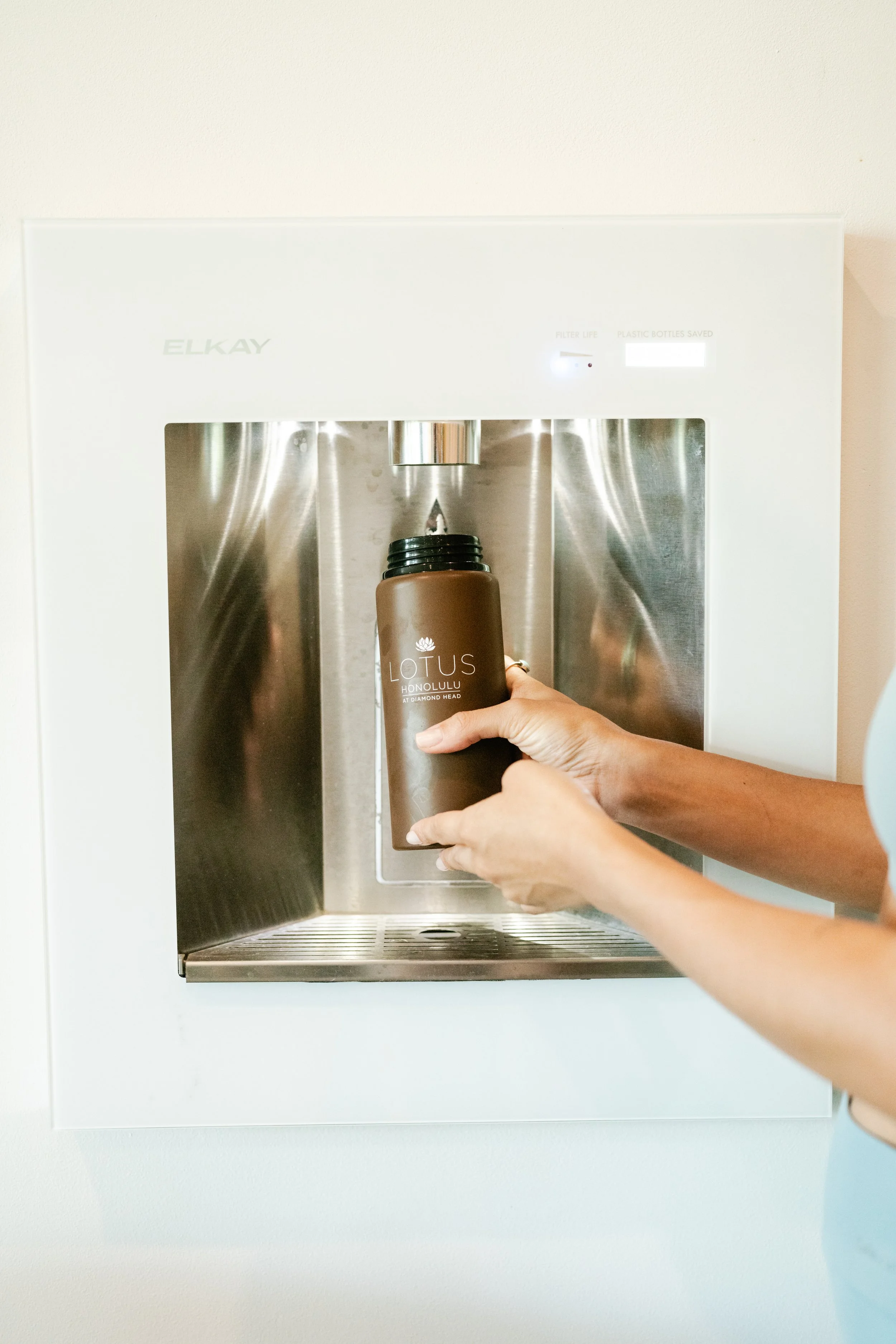 Person filling a brown water bottle labeled 'Lotus Honolulu' at a stainless steel water dispenser.