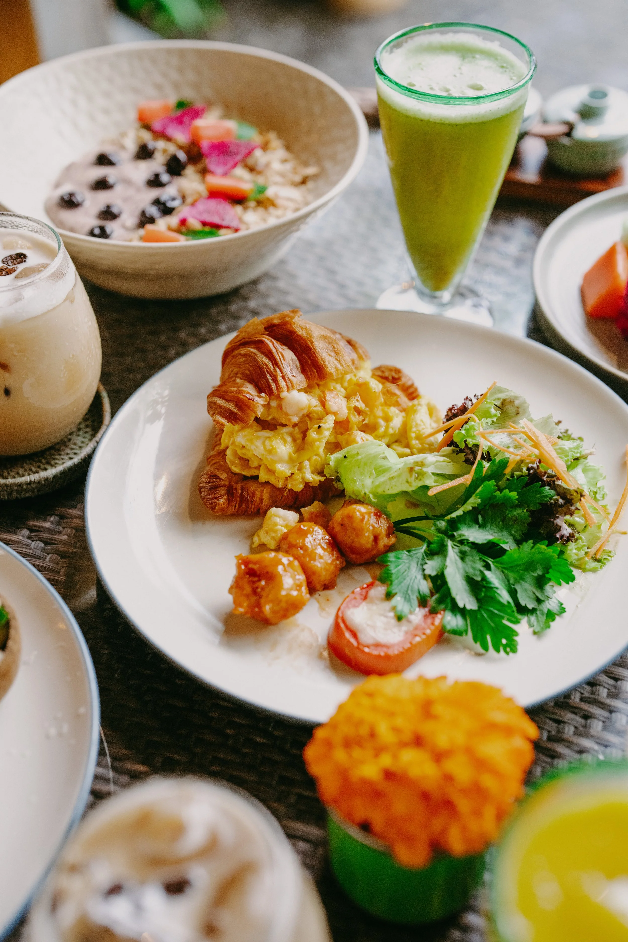 A breakfast spread featuring a plate with scrambled eggs, a croissant, salad, and tomato, with various drinks including a green smoothie, a milkshake, and other beverages, on a table.