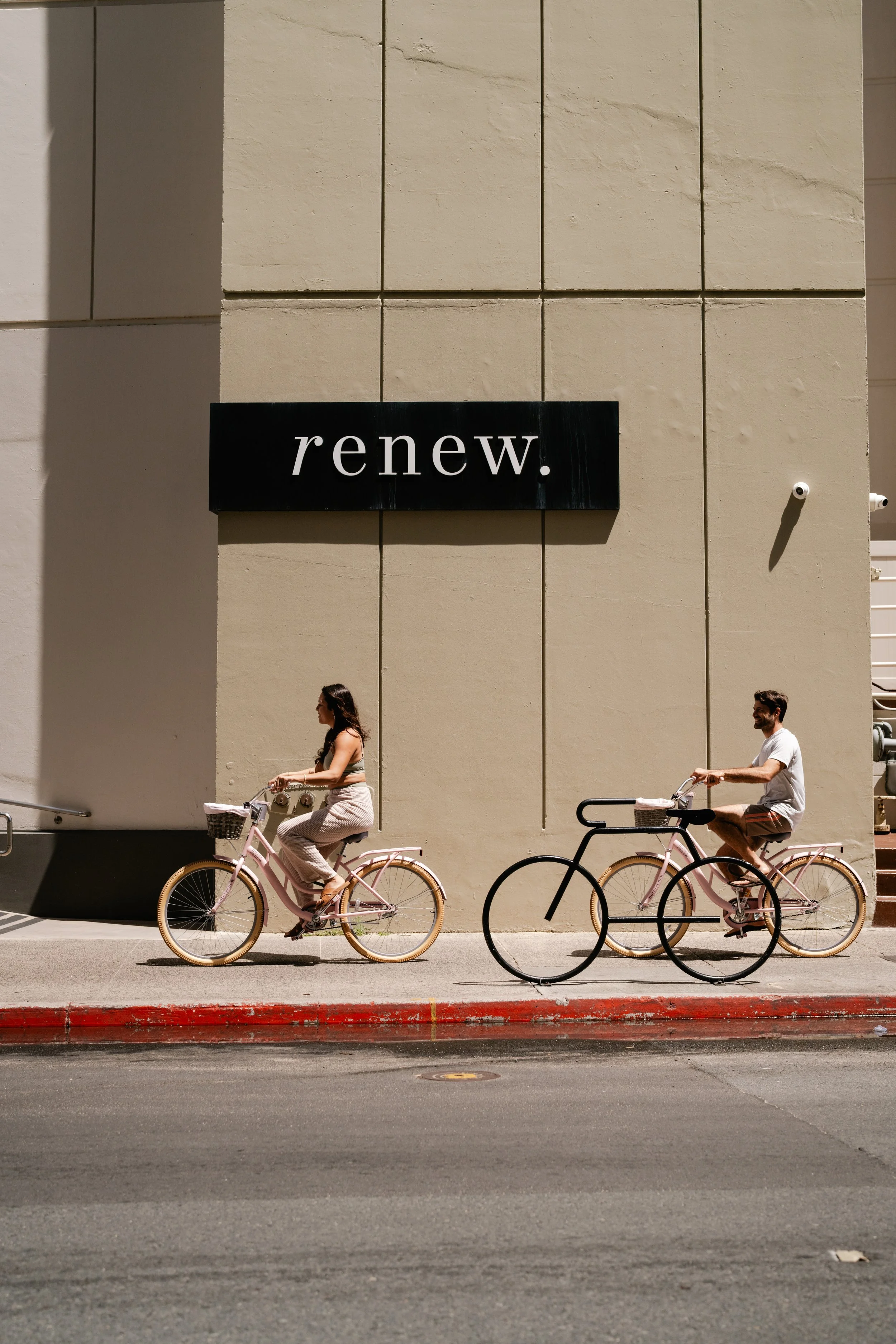 Two people riding bicycles on a city sidewalk beside a beige building with a black sign that reads 'renew.'