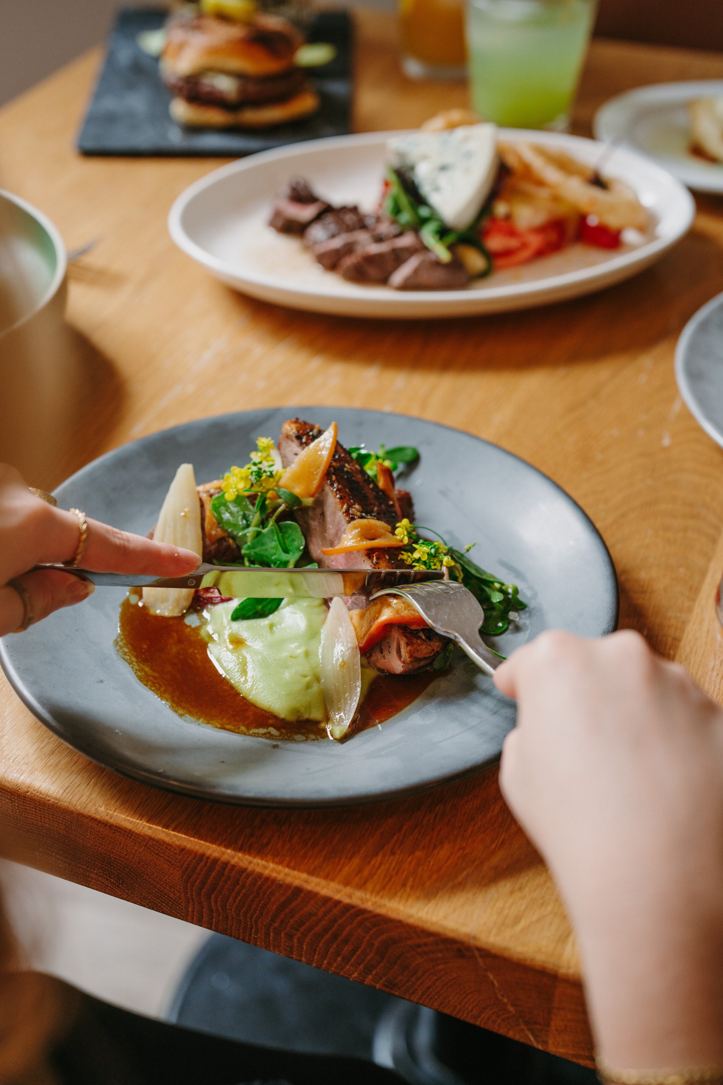 A person cutting into a plated dish of meat with mashed potatoes and vegetables on a wooden table, with other plates of food and drinks in the background.