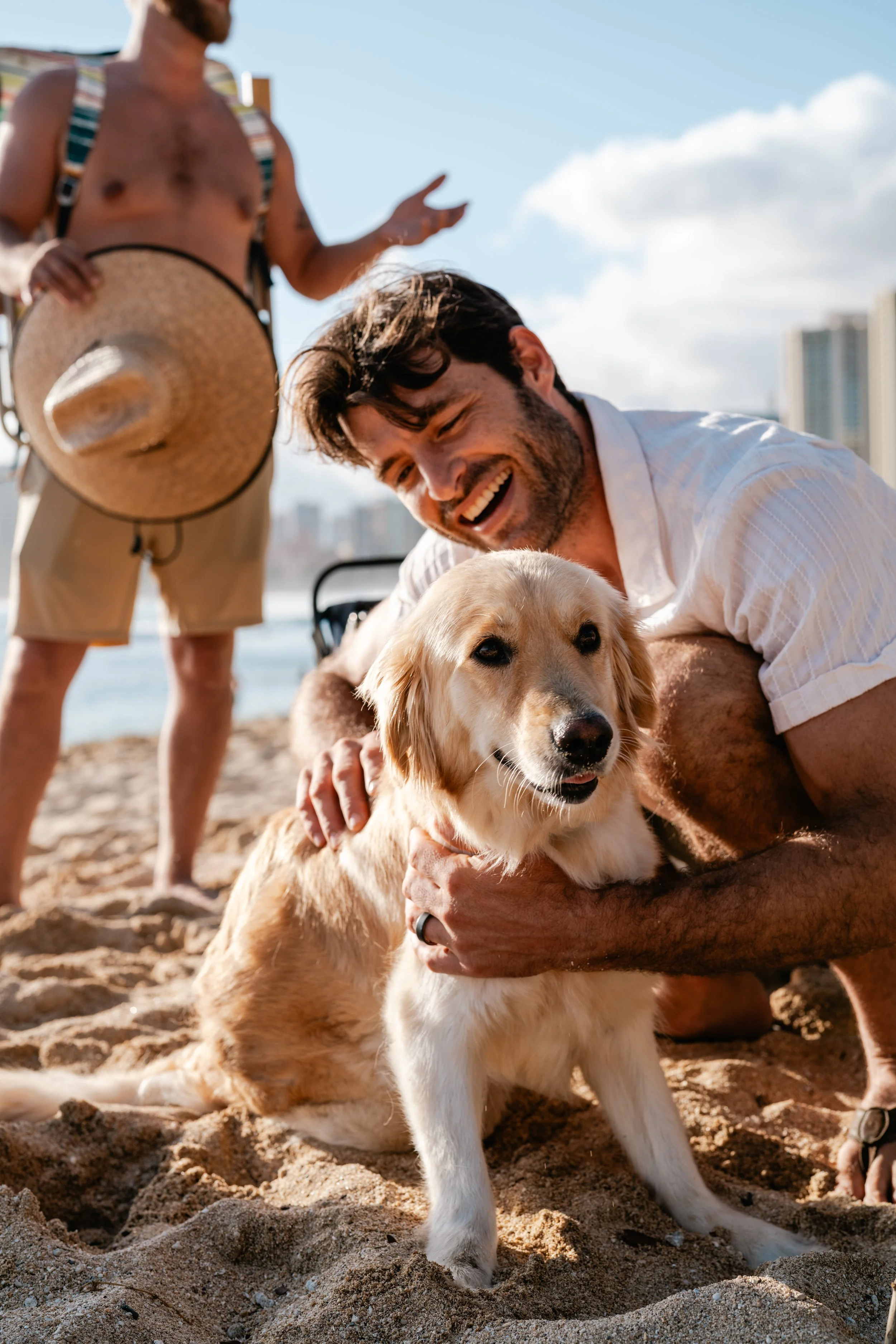 A man and a dog on a sandy beach, with another person in the background holding a straw hat.