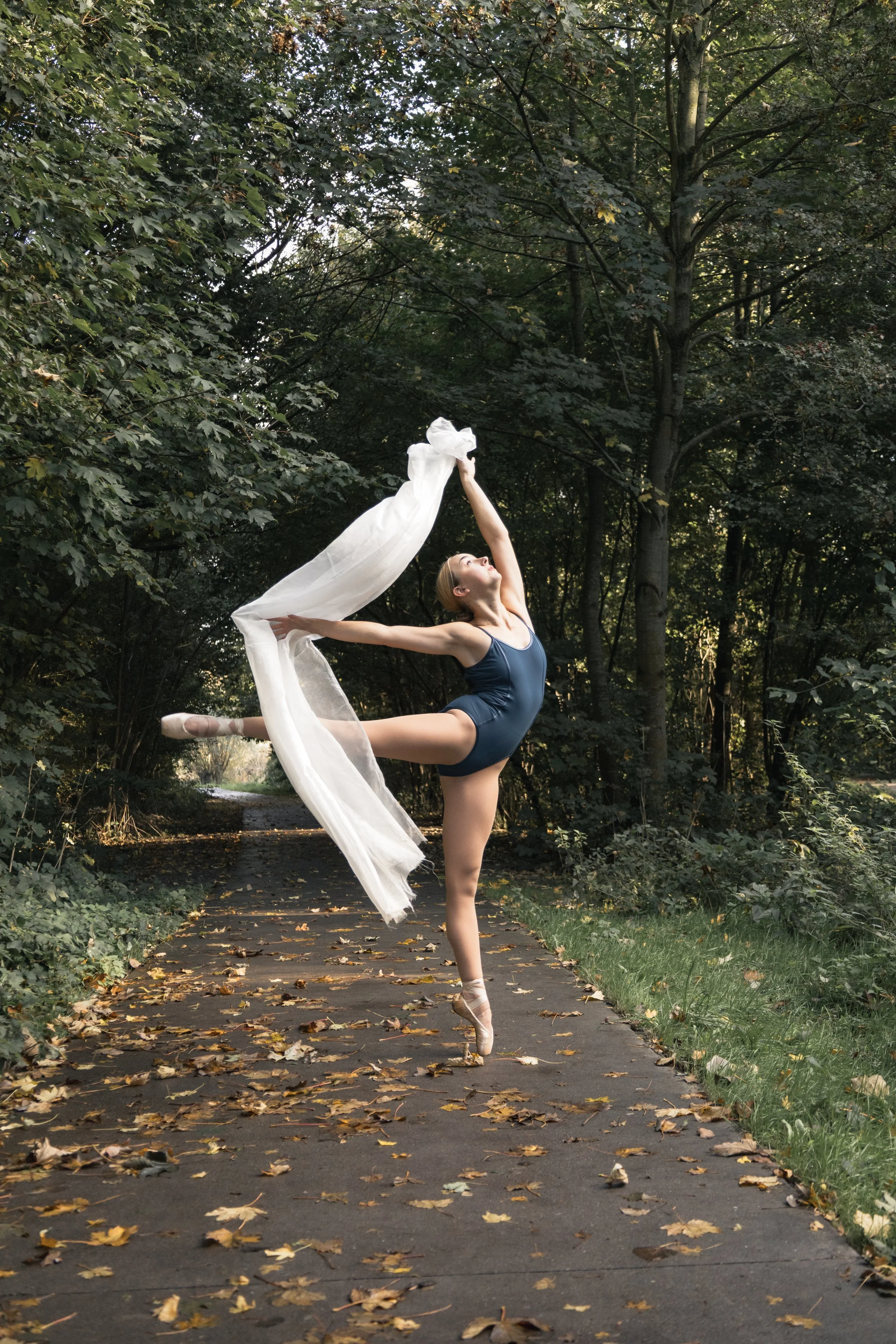 Een balletdanseres in een blauwe balletkostuum danst op een bosweg, haar rechter been in een hoge arabesque en haar linker been op de grond, met een witte doek in haar rechter hand. De omgeving bestaat uit gras, bomen en herfstbladeren op de weg.