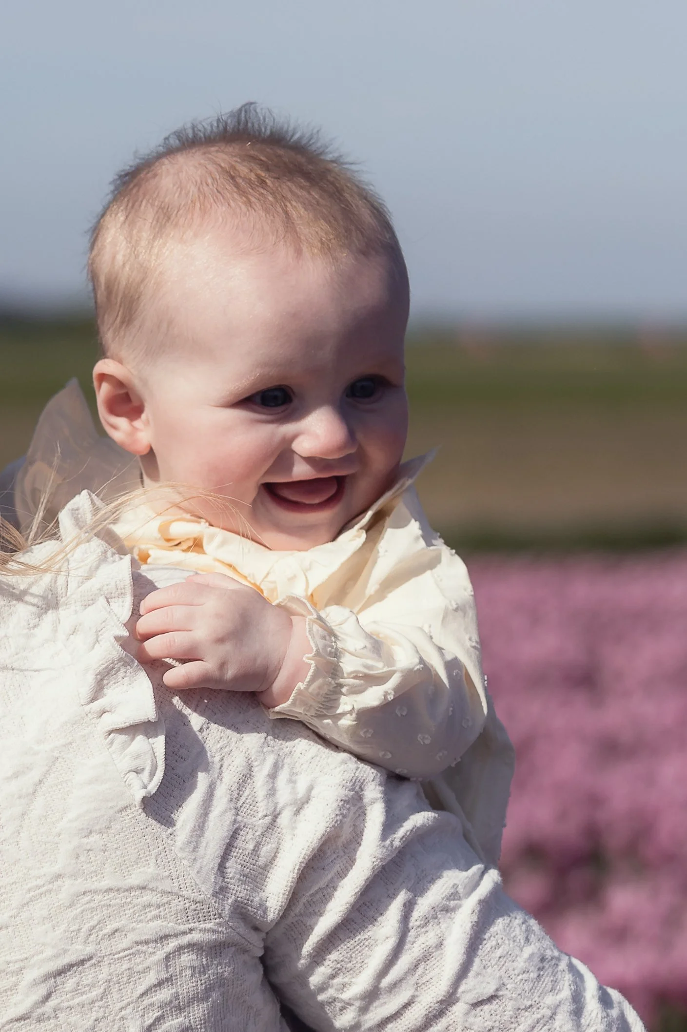 Close-up van een lachende baby met blauwe ogen, in een crèmekleurige jas, terwijl hij in een veld staat met bloemen onder een heldere blauwe hemel.