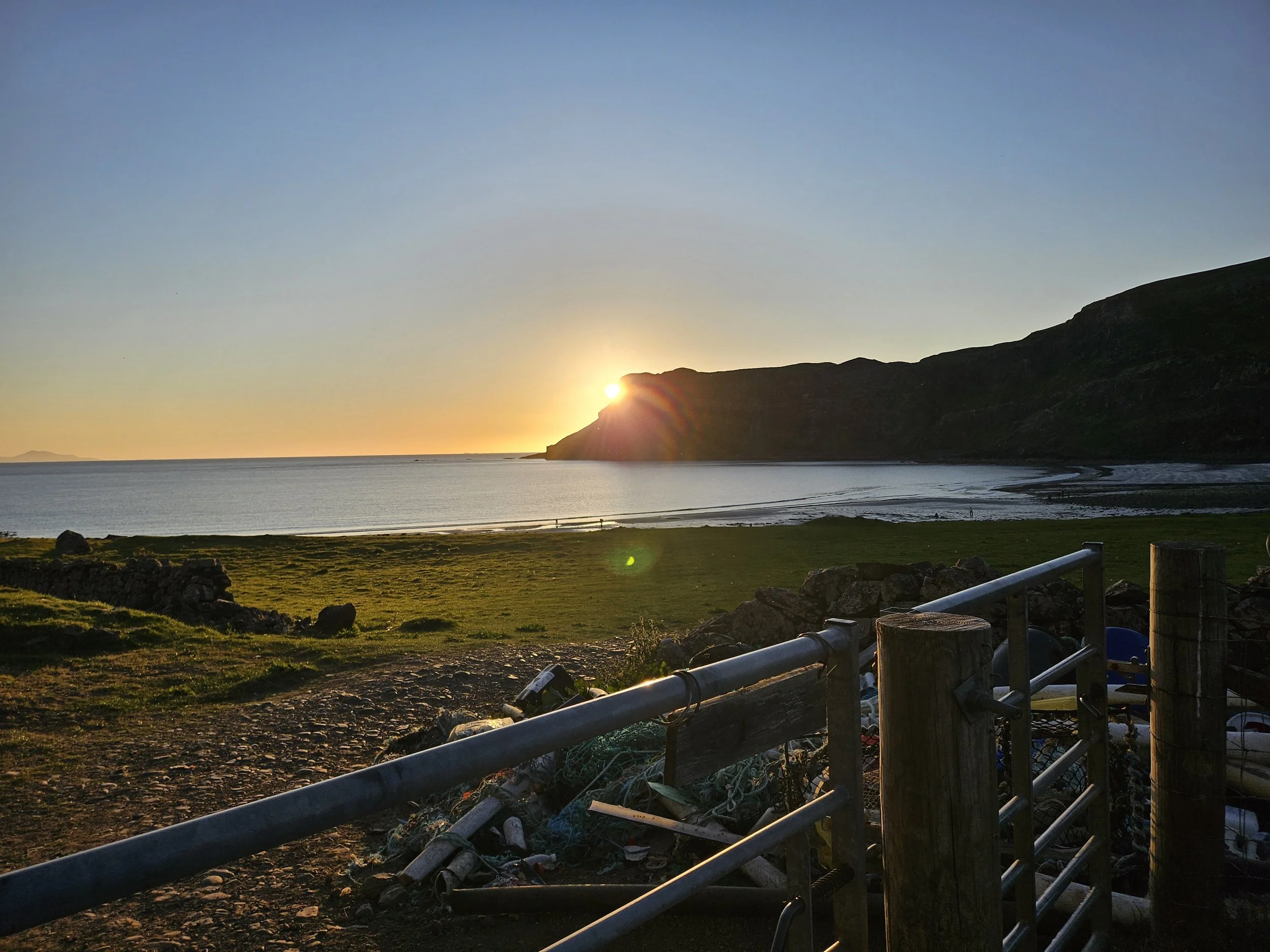 Sunset over a grassy coastline with hills in the background and a metal fence in the foreground.