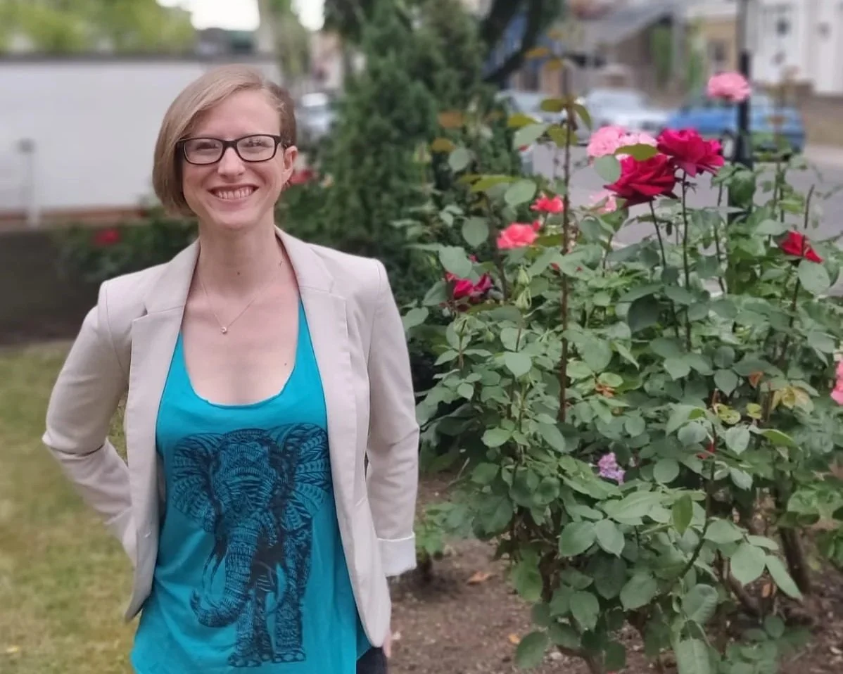 Smiling woman with short blond hair, glasses, and a light blazer, standing outdoors near a bush of pink and red roses, in a residential neighborhood with houses and cars in the background.