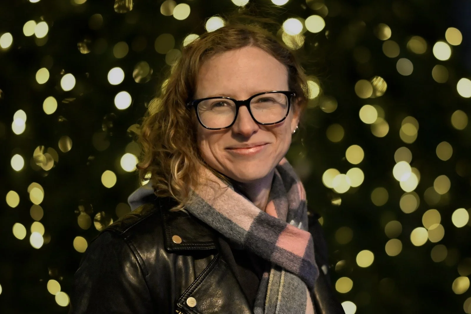 A woman with curly hair and glasses smiling at the camera, wearing a leather jacket and a plaid scarf, with blurred yellow holiday lights in the background.