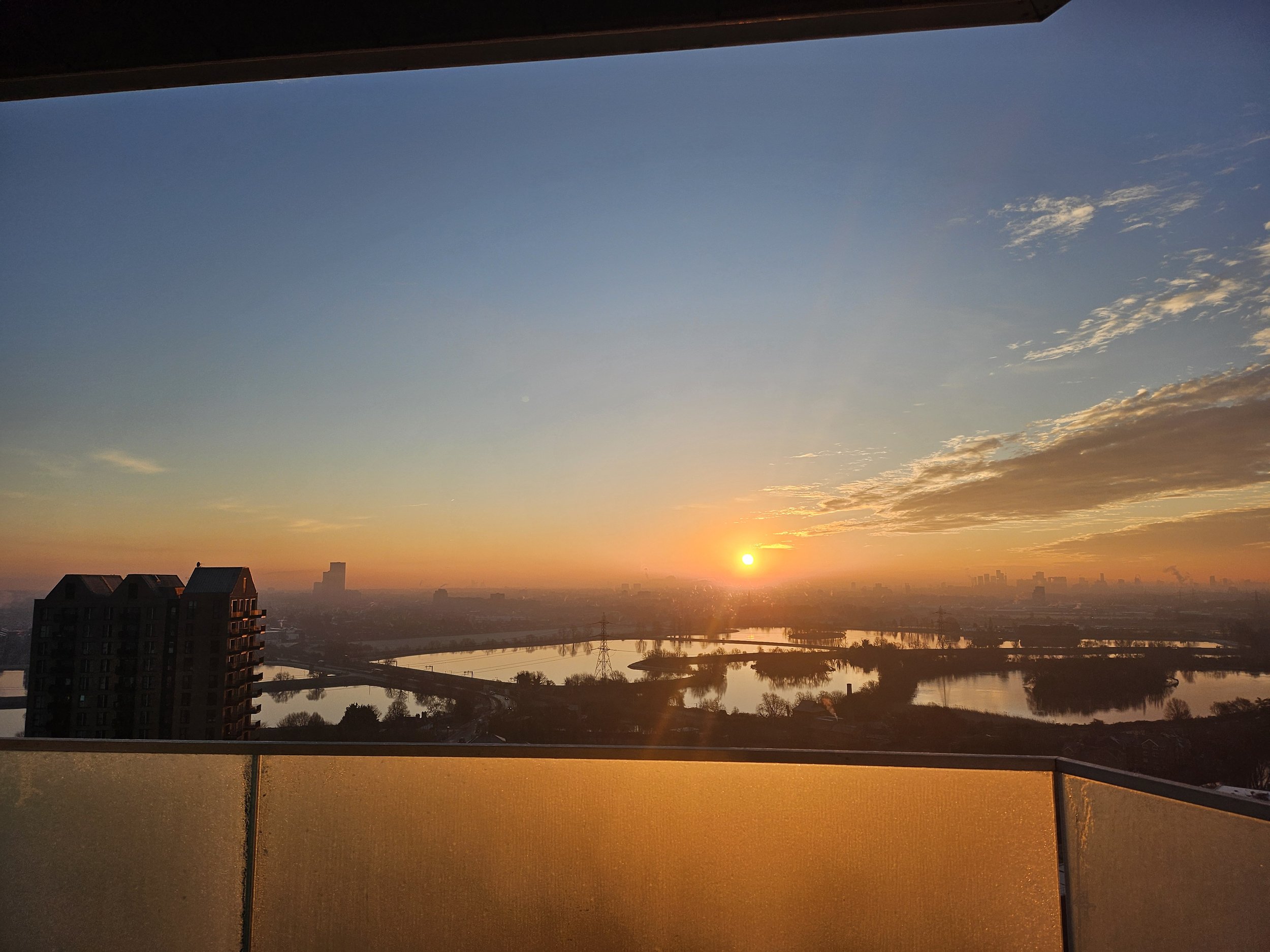Sunset over a city with water bodies, buildings, and a bridge, viewed from a high-rise balcony with a glass railing.