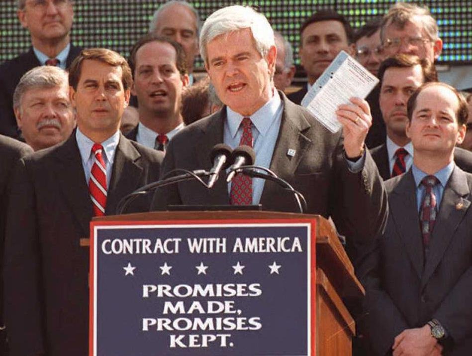 A man with gray hair speaking at a podium during a political rally, holding a paper in his hand. He is surrounded by several men in suits, some with striped ties, on a stage with a sign that reads "Contract with America: Promises Made, Promises Kept." in front of the podium.
