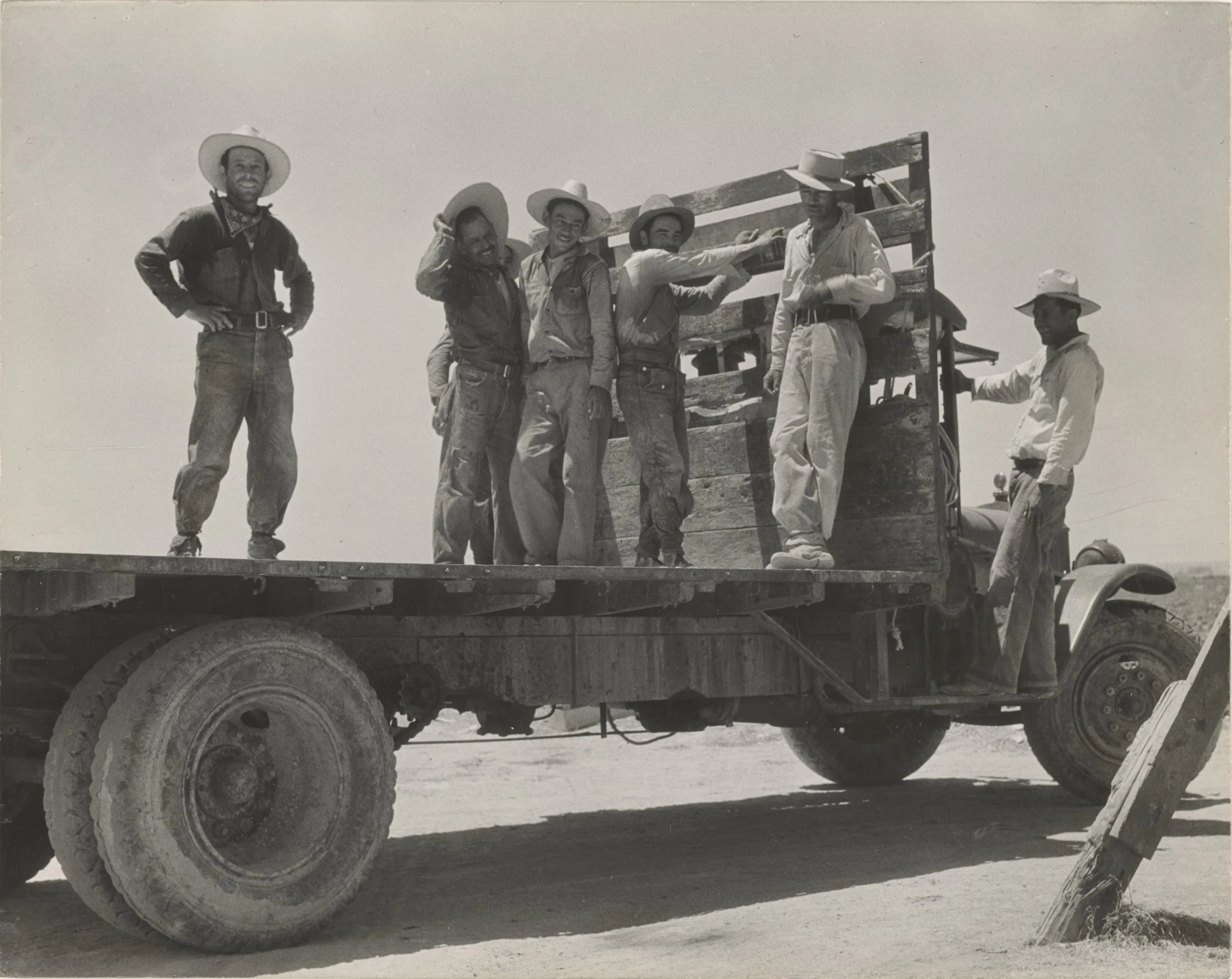 Black and white photo of seven men with hats standing on a flatbed truck in a desert. Four men are on the truck bed, and two are near the cab. The background shows a clear sky and open land.