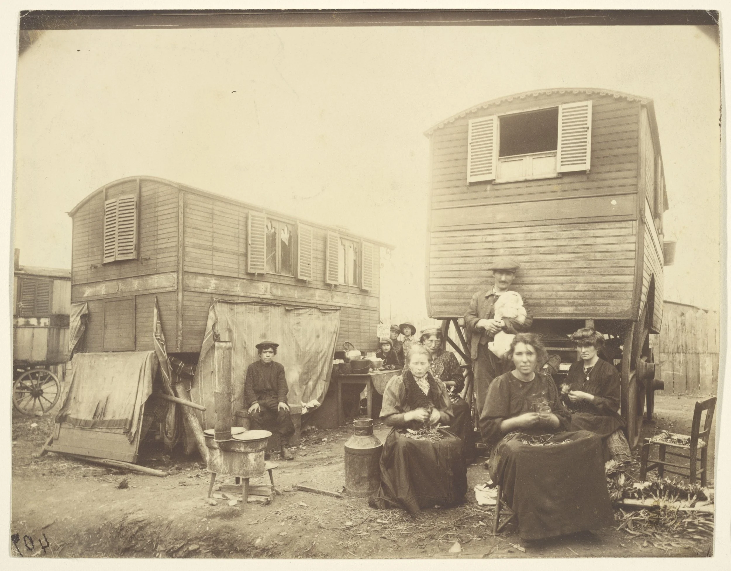 A vintage black-and-white photograph showing a group of women, a man, and children gathered outdoors in front of wooden house trailers during the early 20th century. The women are seated, some holding jars or items, and a woman stands holding a child
