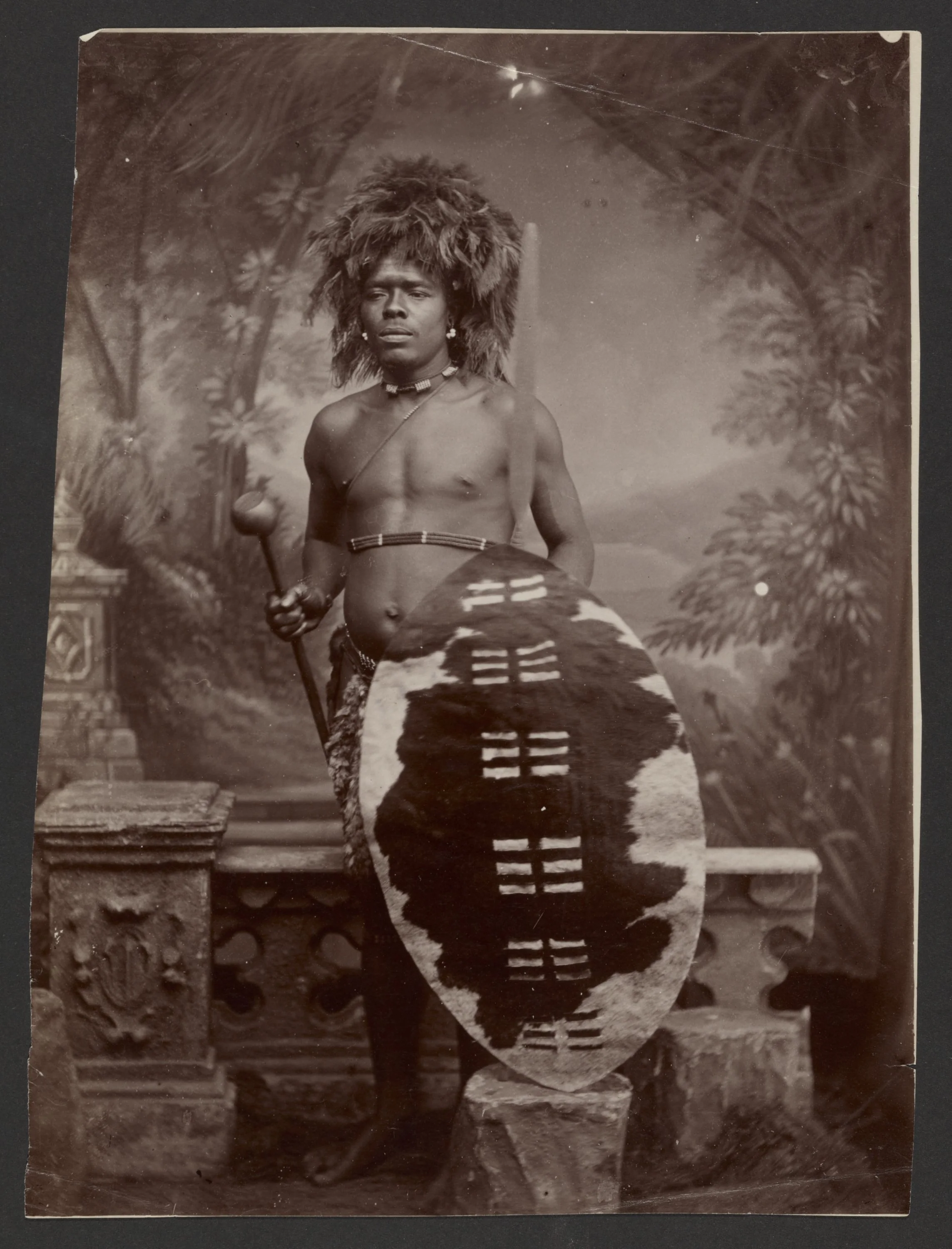A man dressed in traditional African attire holding a spear and shield, standing in front of a painted backdrop depicting a natural landscape with trees.
