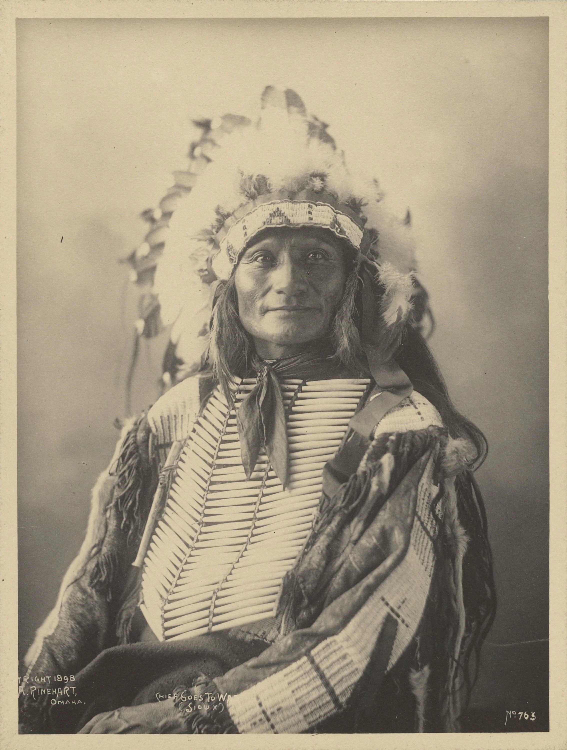 A black and white portrait of a Native American man wearing a traditional feathered headdress and clothing, looking directly at the camera.