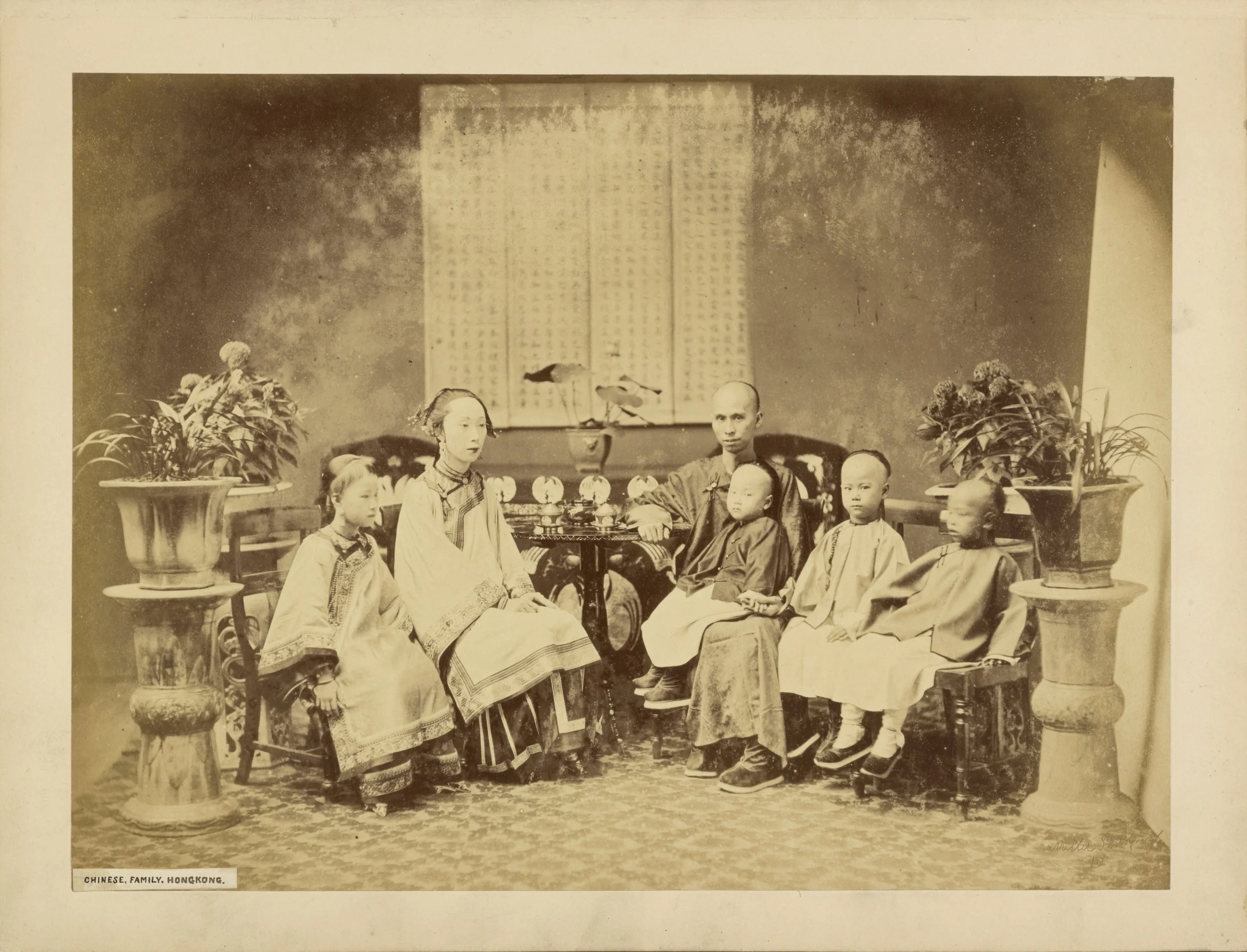 A vintage sepia photograph of a Chinese family posing indoors, with four children and two adults, around a table with potted plants and a decorative background.