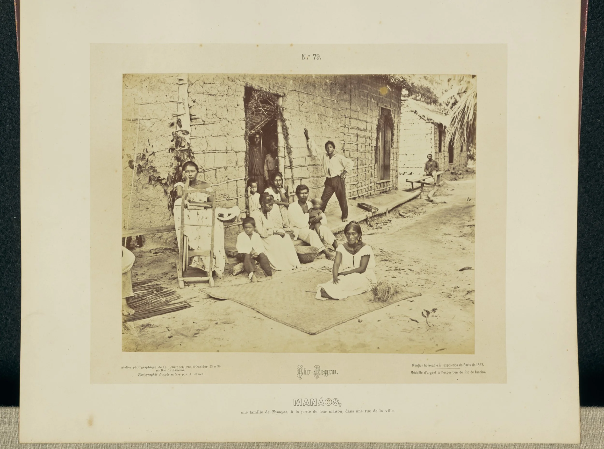 A vintage black and white photograph of a family sitting on steps outside a rustic house in Manaus, Brazil, with some children seated on a mat on the ground in front. The house appears to be made of mud bricks, and the surrounding area is unpaved.