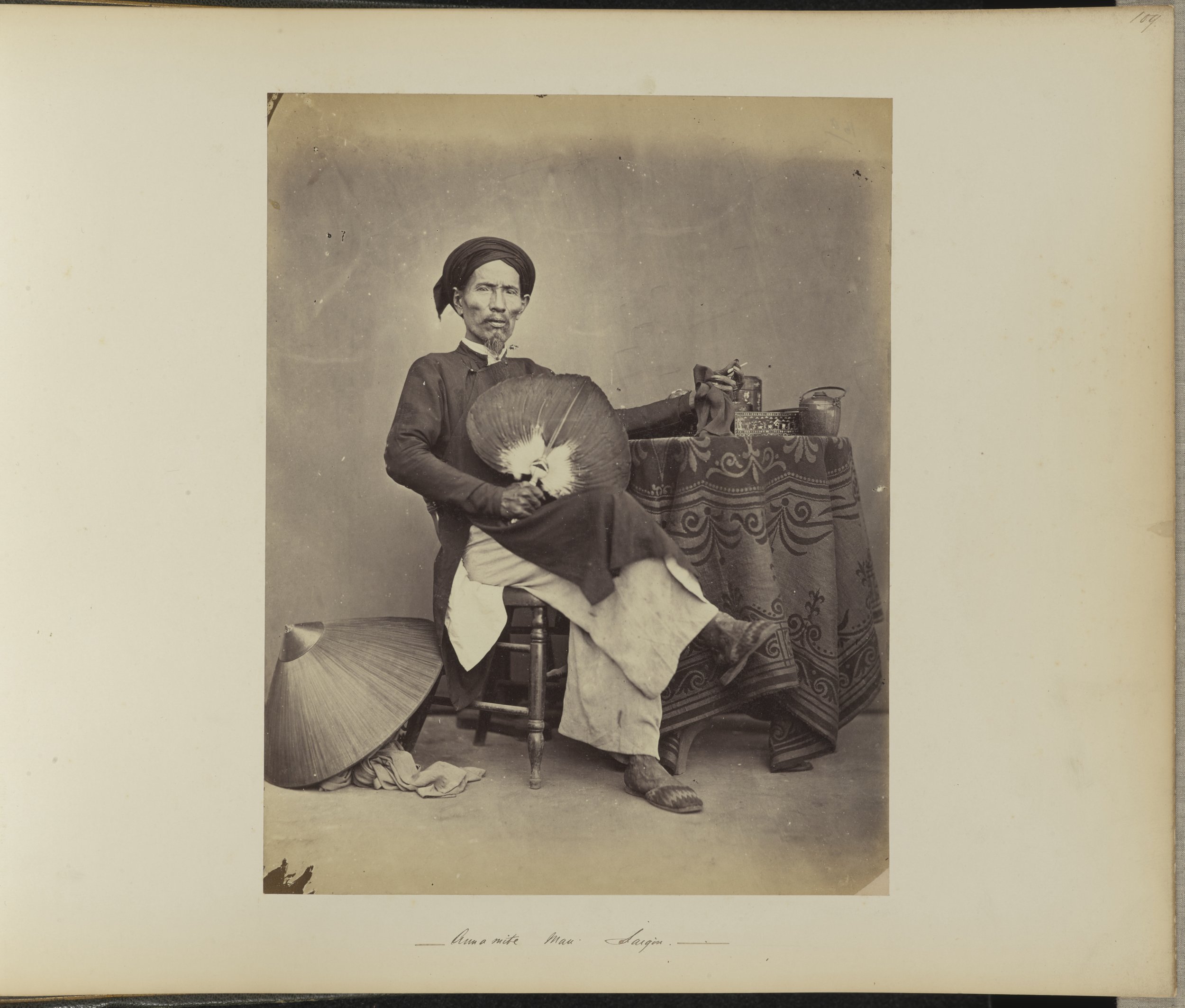 An Asian man sitting on a stool, holding a fan, wearing traditional clothing and a head covering, with a parasol on the floor beside him, and a table with decorative items in the background.