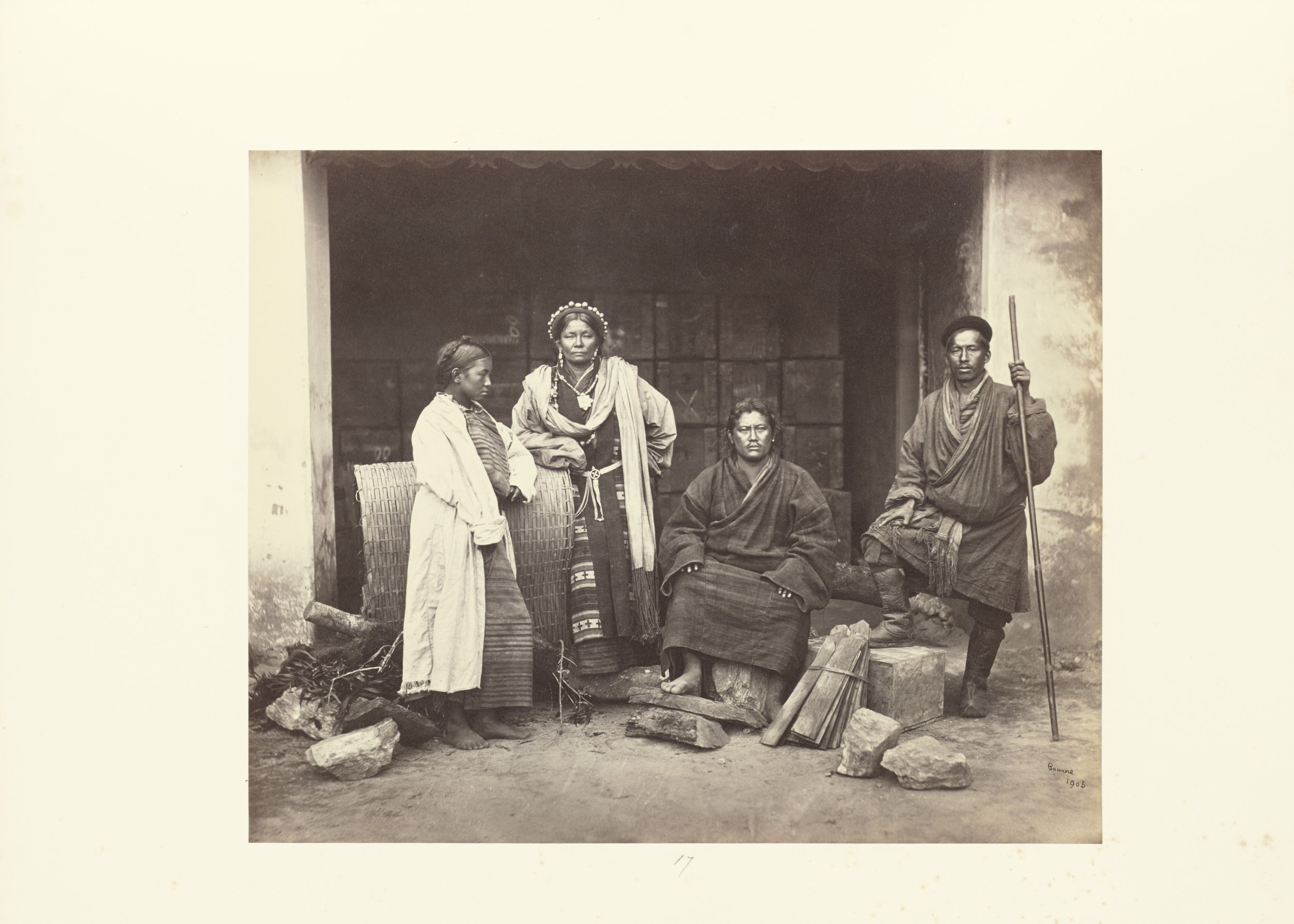 Black and white photograph of four indigenous people, three women and one man, standing and sitting outside a structure with a doorway. The woman on the far left is looking down, wearing a white outer garment with a striped skirt. The second woman fr