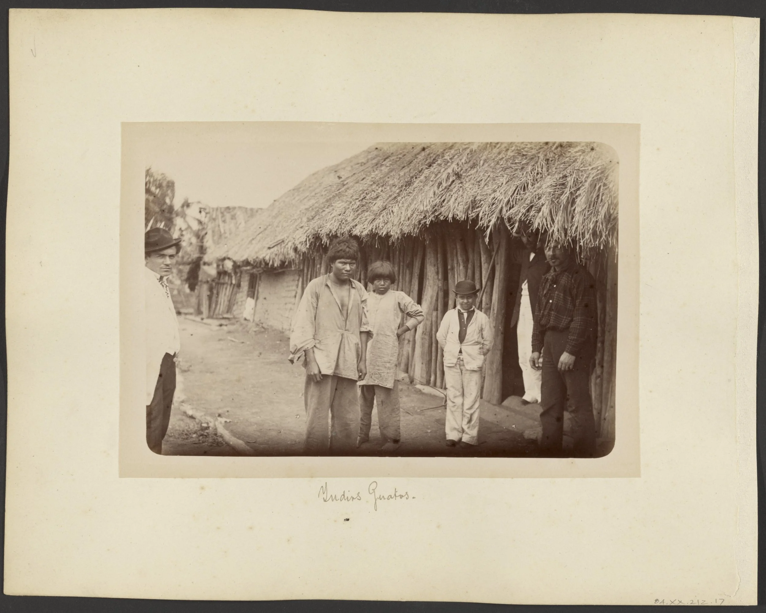 Vintage black and white photograph of five people standing in front of a thatched roof hut, with two men on the left and right, and three children in the middle, outdoors in a rural setting. The caption reads "Undips Guatos."