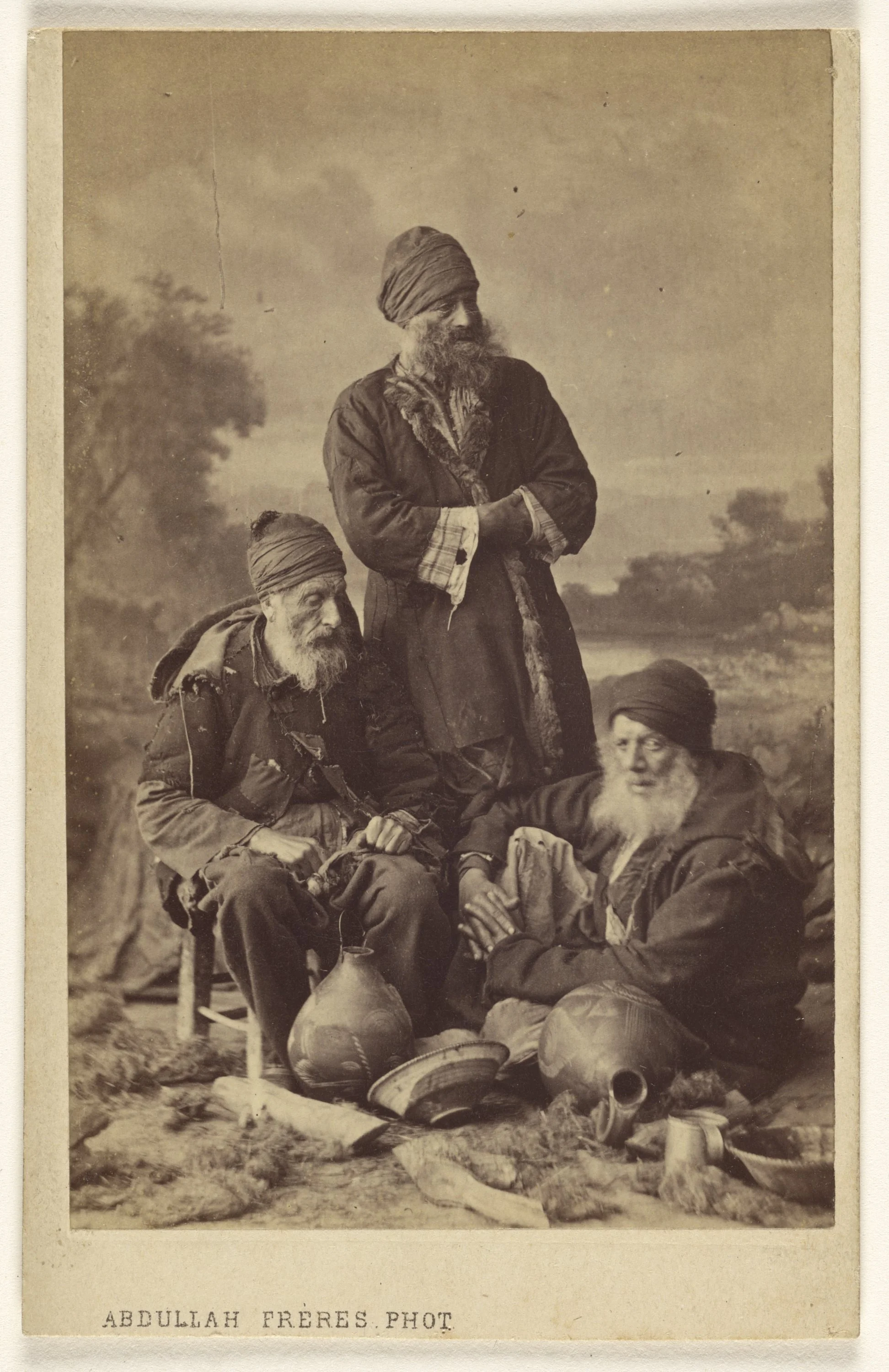 Historical photograph of three elderly men with beards and traditional clothing, placed outdoors with trees in the background. Two of them are sitting; one on the ground leaning on his side, and the other on a chair, while the third man stands behind