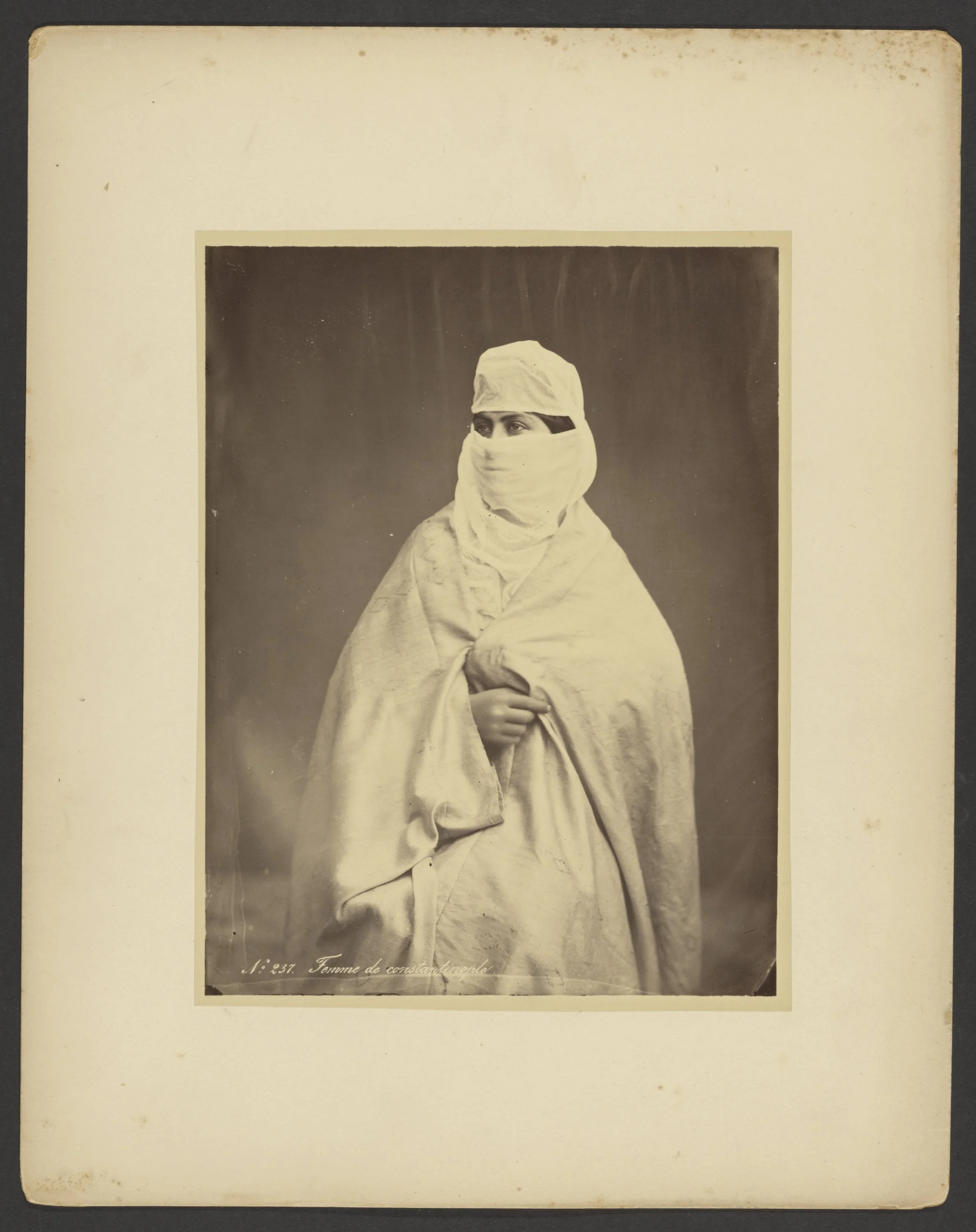 A black-and-white historical photograph of a woman dressed in traditional Islamic attire, including a head covering and face covering, with only her eyes visible, standing against a plain background.