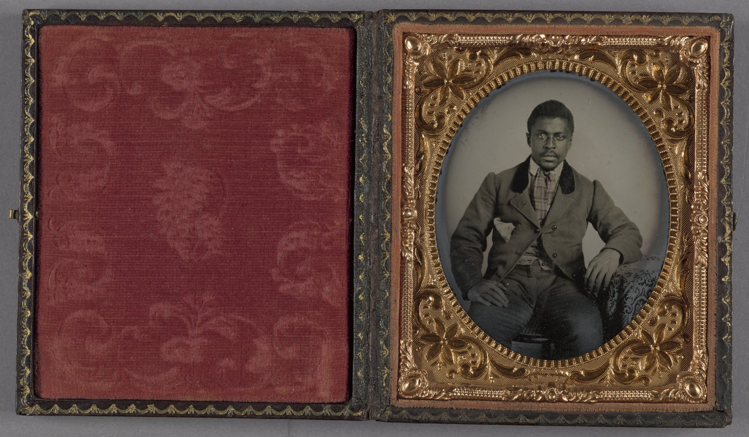 A vintage black-and-white portrait photograph of a man, seated with one hand on his knee, in an ornate gold frame within a red velvet-lined case.