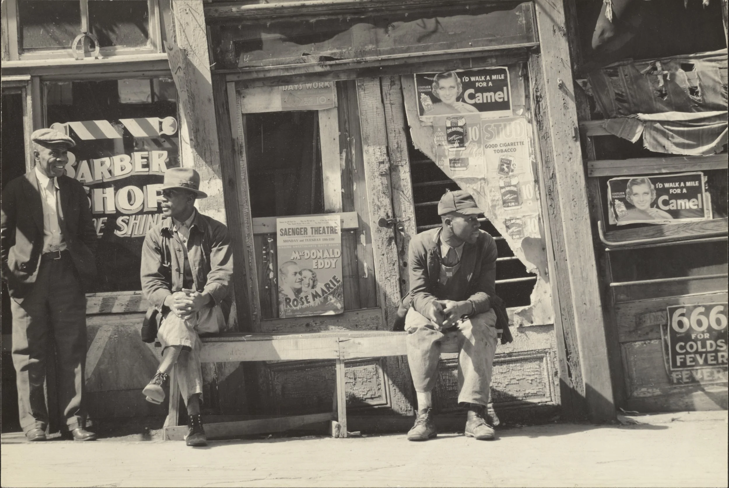 Three men sitting and standing outside a weathered storefront on a city street. The storefront has posters and signs, including one for a theater showing comedians McDonald Eddy and Rose Marie, and others advertising cigarettes and cold remedies. Two