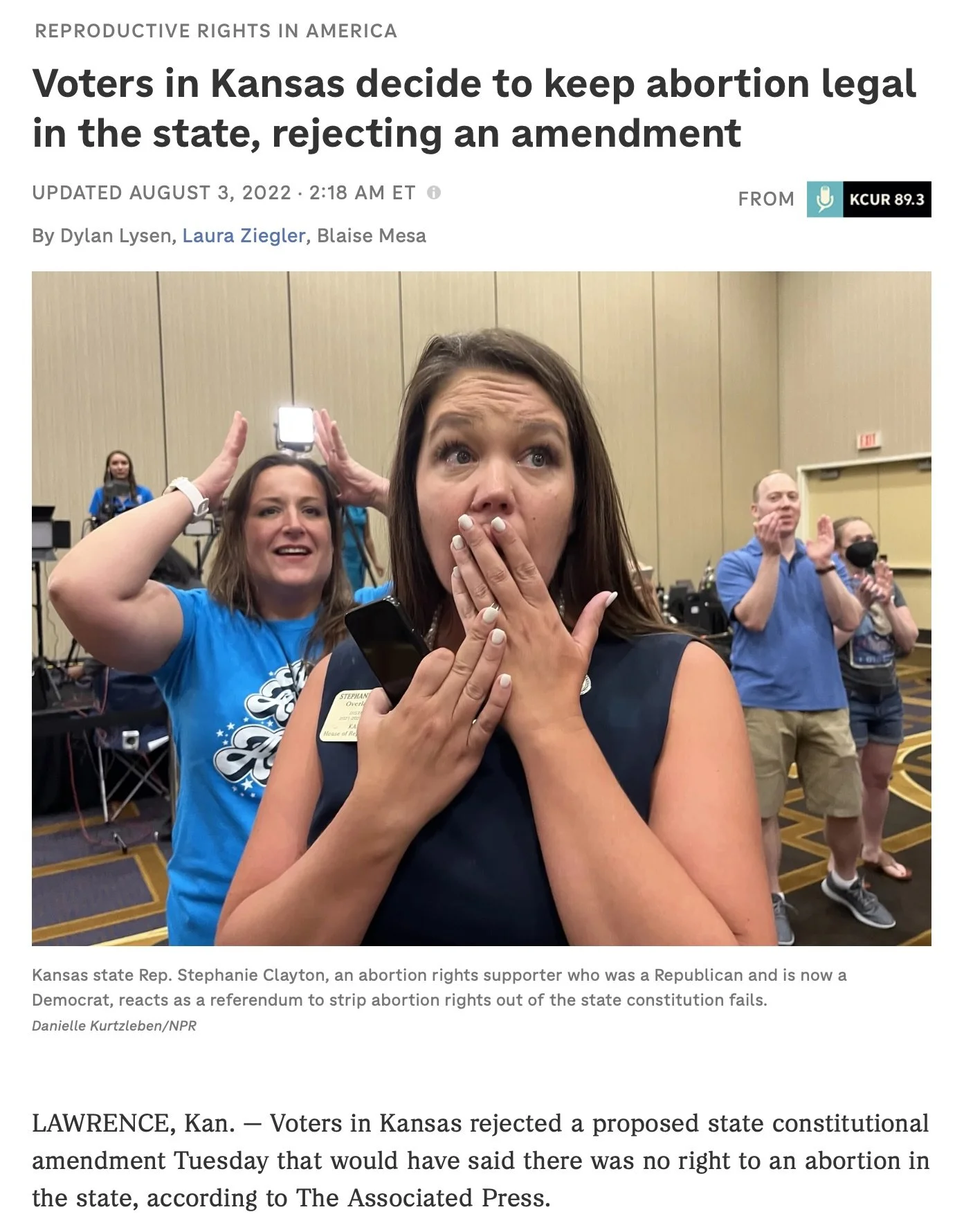 A woman with long brown hair and a sleeveless top reacts with surprise, covering her mouth with one hand and holding a phone in the other. Behind her, a woman in a blue shirt and a man in a blue polo shirt are clapping, and other people are standing in a room with beige walls, some wearing masks.
