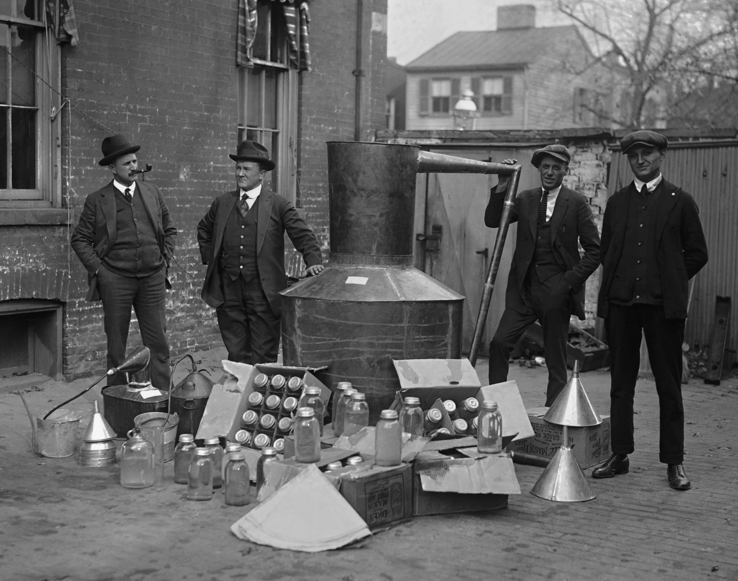 Four men in vintage suits and hats standing outside near a large industrial machine, surrounded by glass jars, boxes, and metal cones.