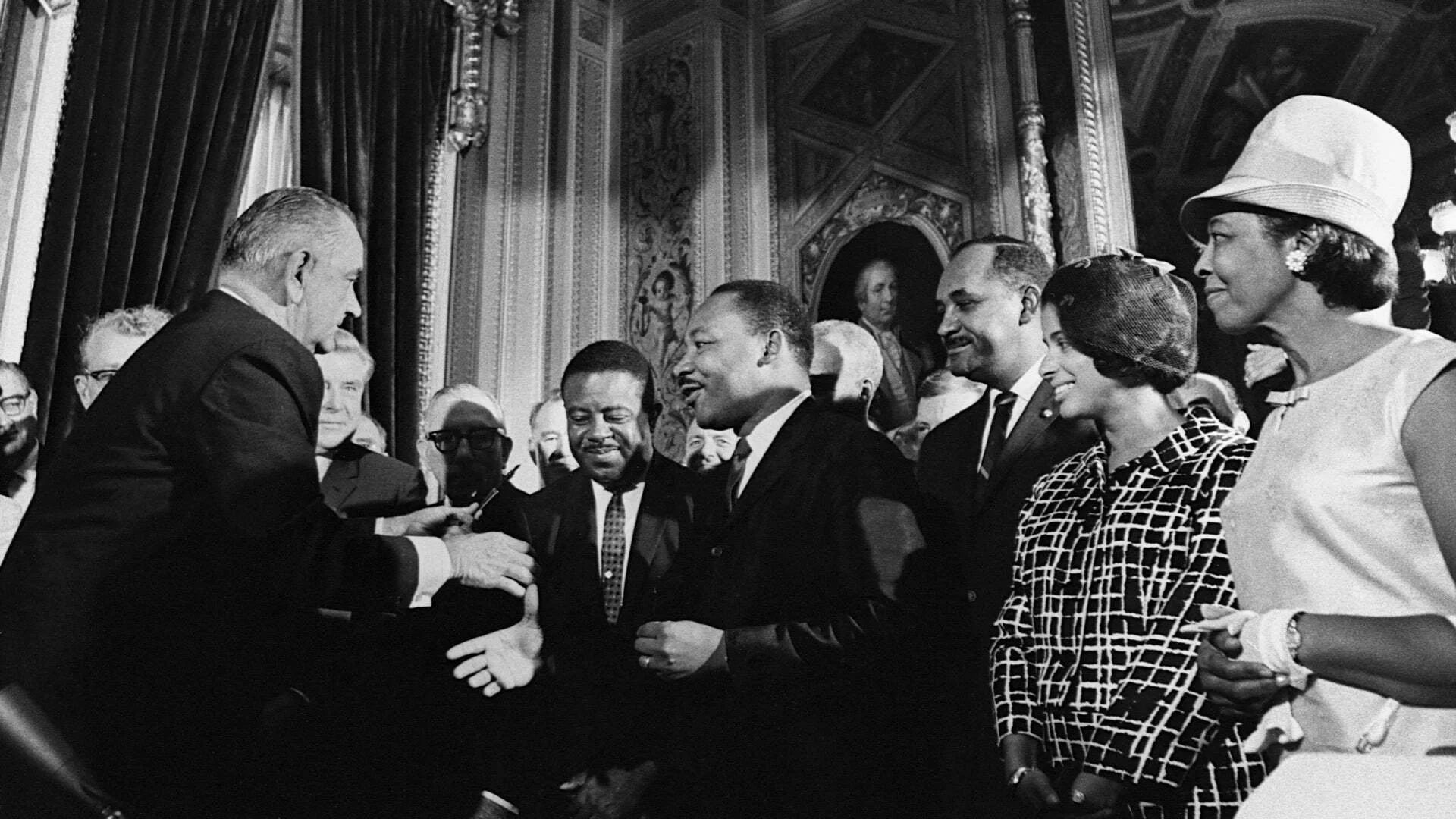 A black and white photograph of a formal event with several people standing in a line, including a man with a beard, a woman wearing a hat, and a man of color, engaging in conversation.