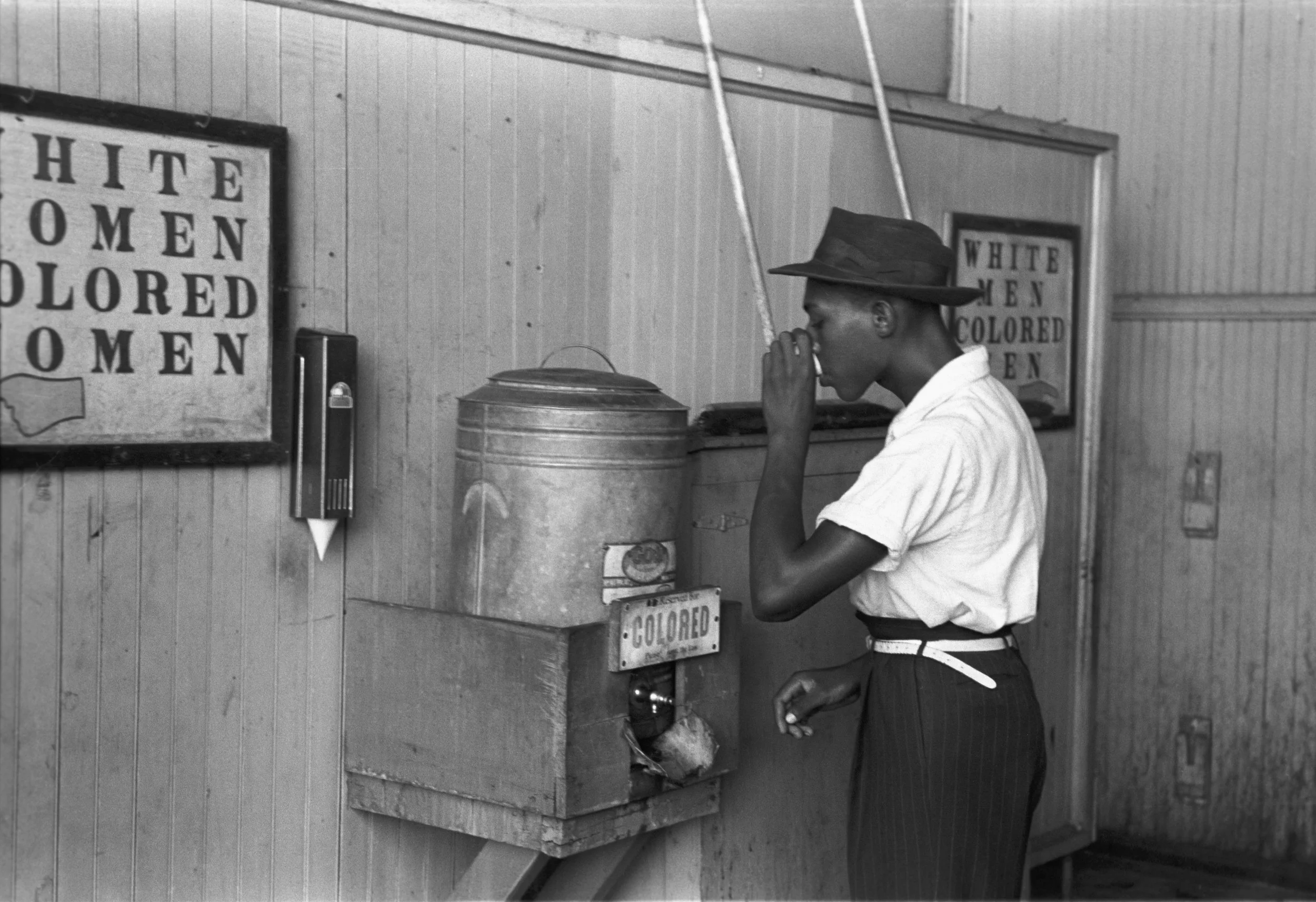 A young African American man wearing a hat, white shirt, and pinstriped pants, standing at a soda fountain labeled 'Colored,' and whispering into his hand in a segregated setting with signs indicating racial separation.