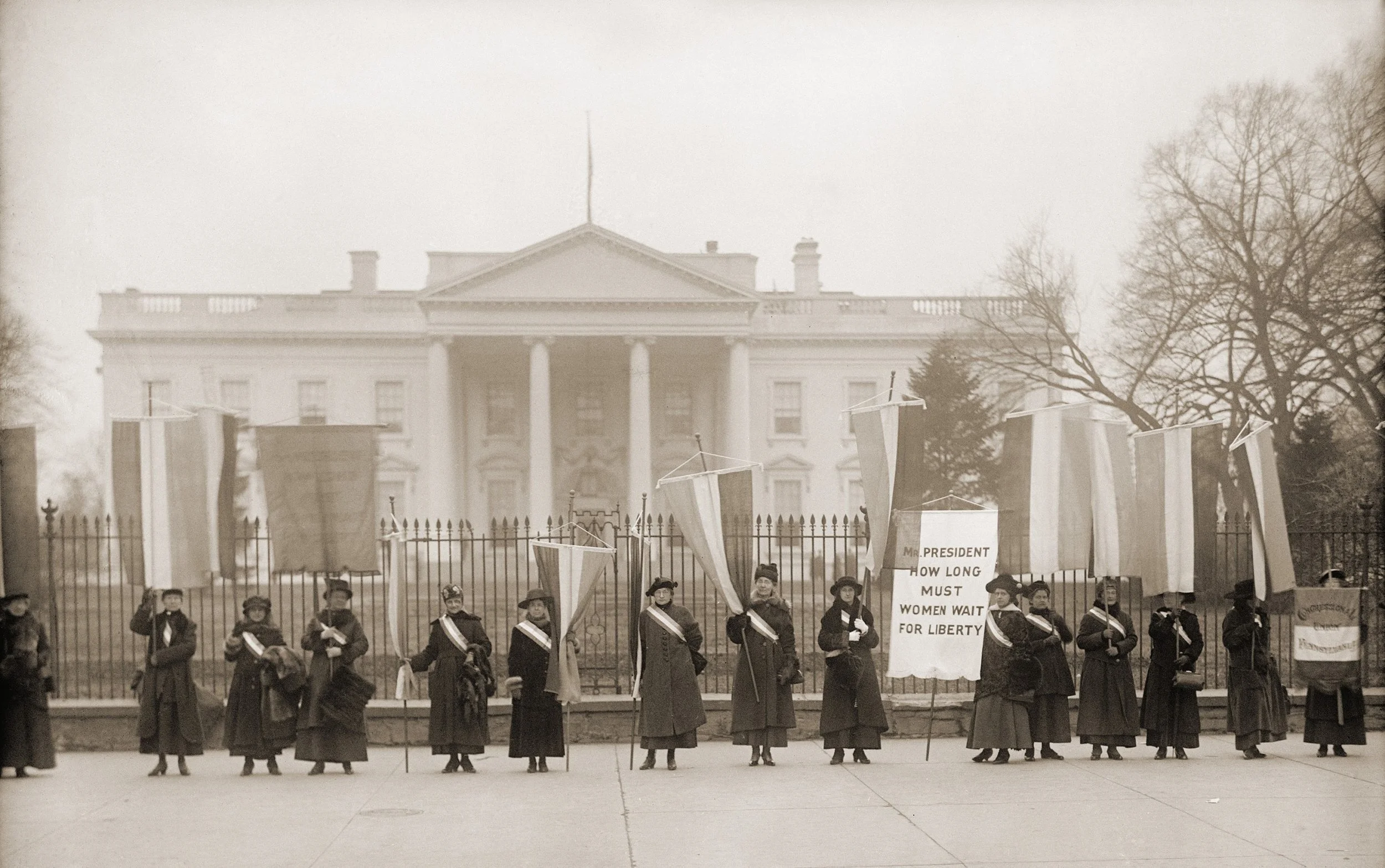 Women protesting outside the White House holding signs and banners advocating for women's suffrage and voting rights.